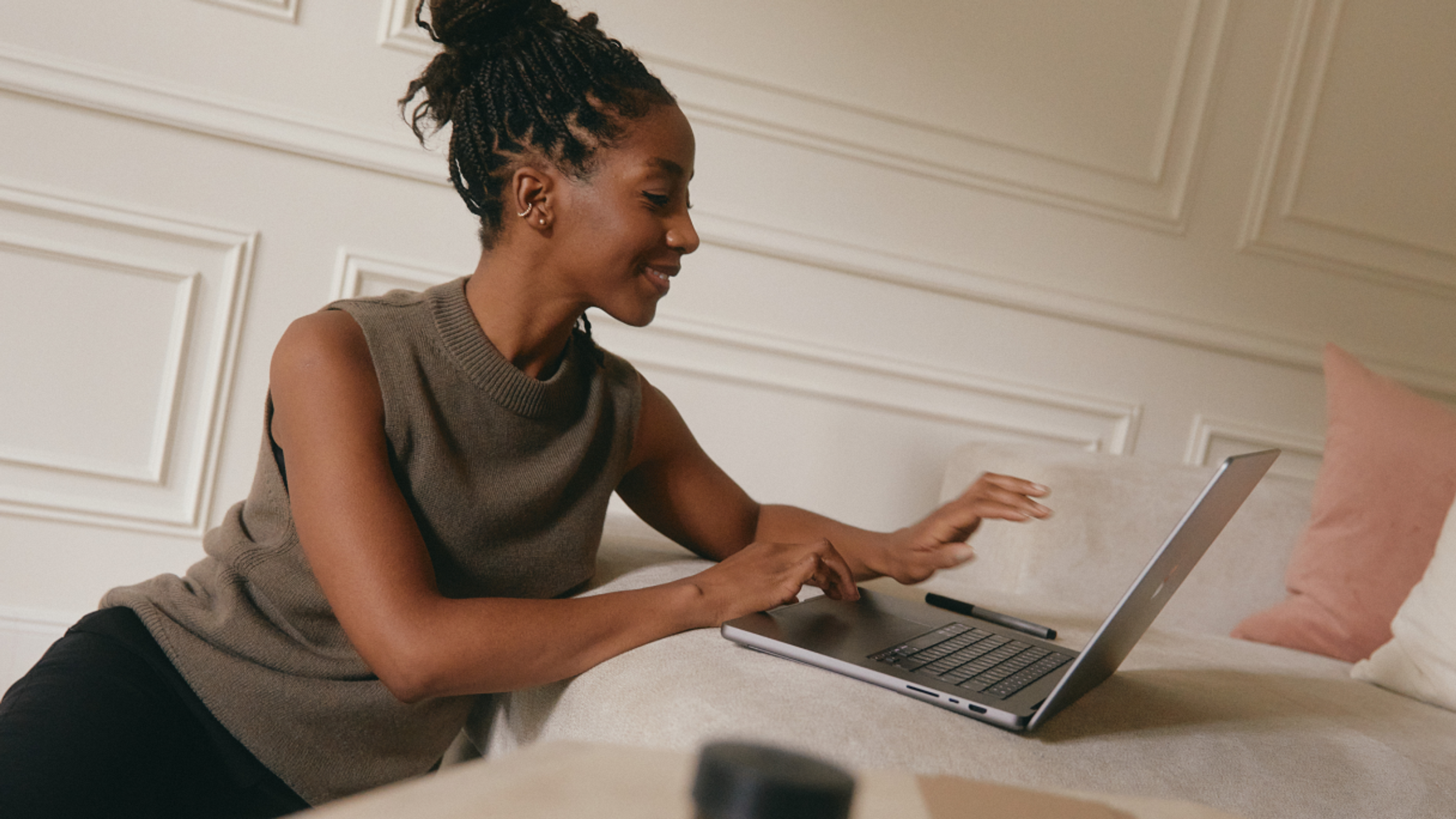 A woman sits on a light sofa, smiling as she works on a laptop in a stylish, softly lit room with white paneled walls and pink cushions.