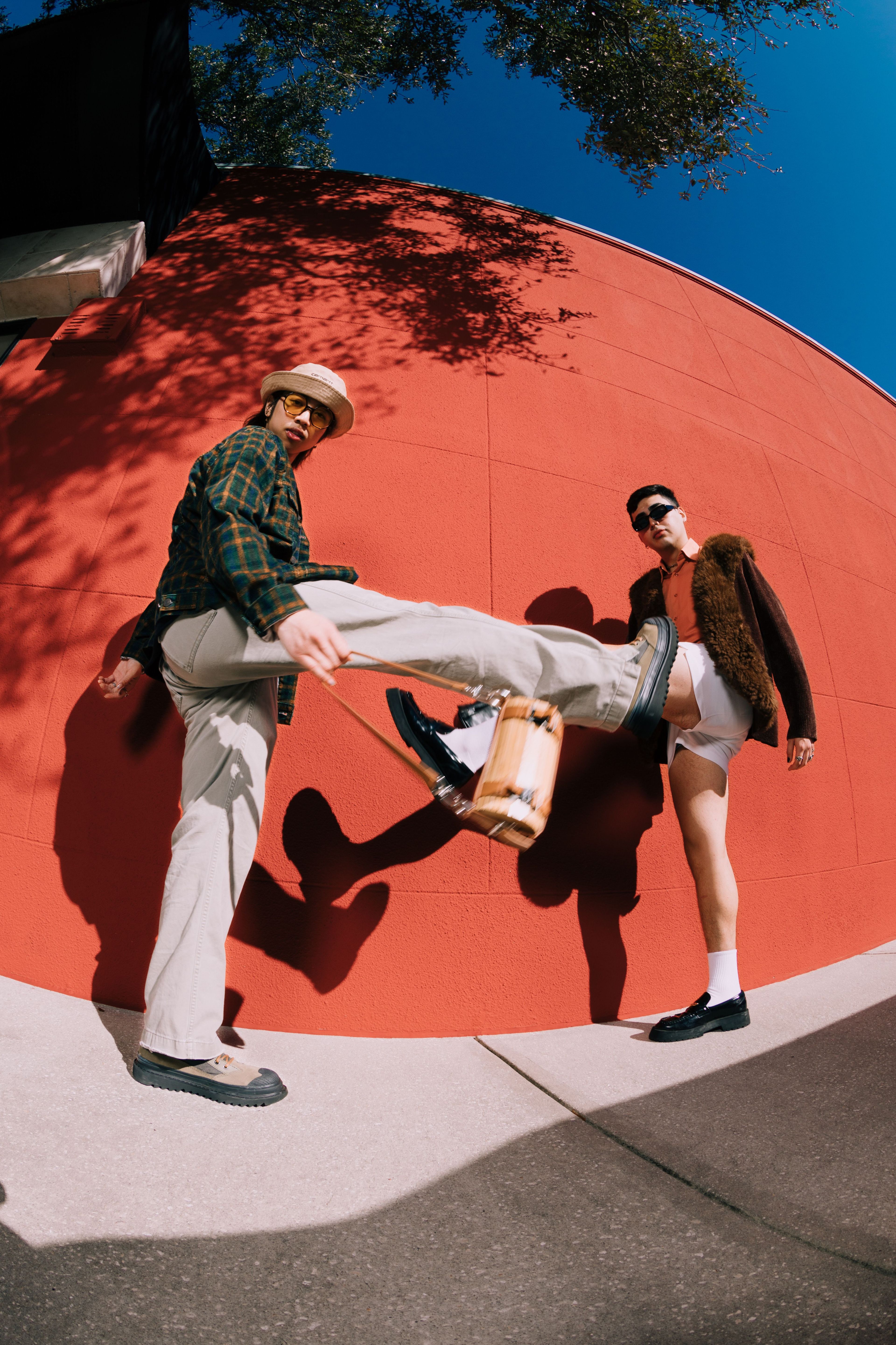 Two stylish men pose outdoors against a bright red wall. One man in light pants and a hat lifts his leg toward the camera, while the other, in shorts and sunglasses, stands confidently beside him.