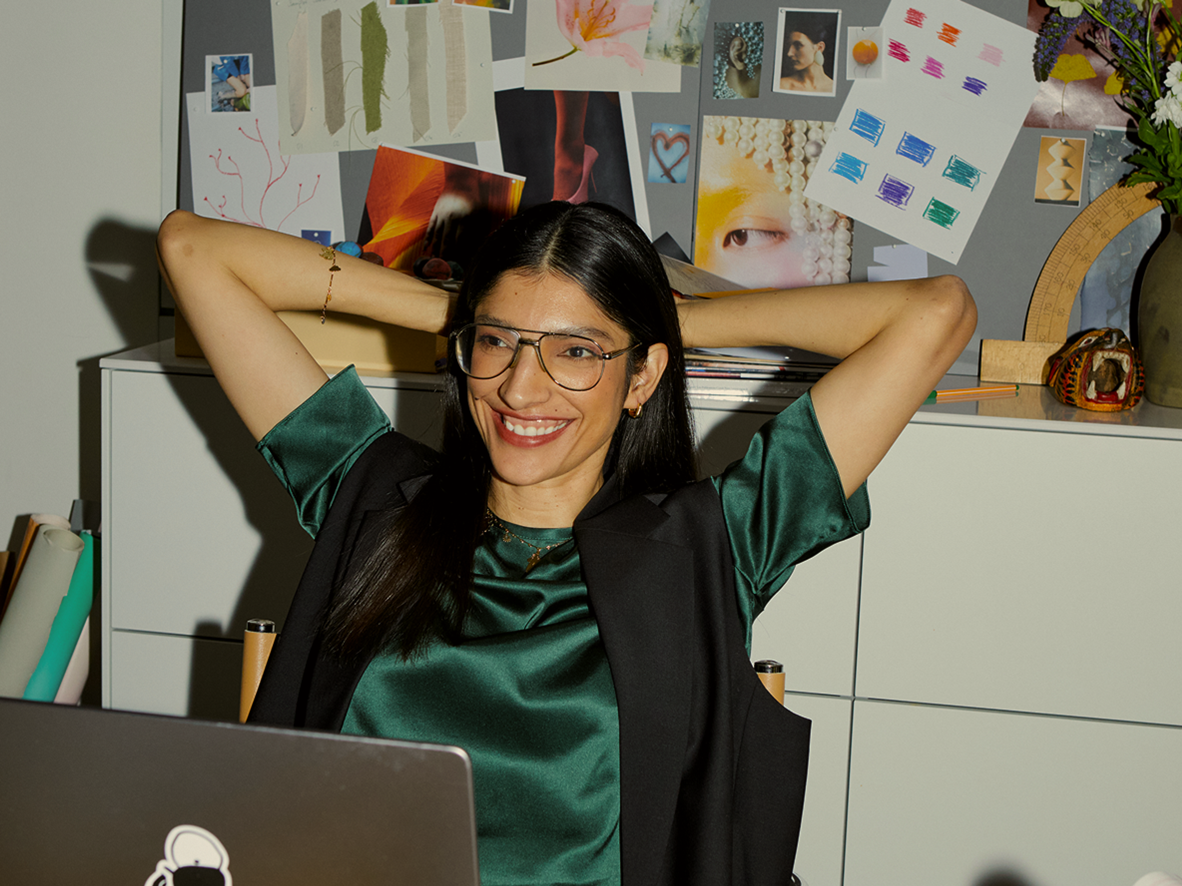 A woman with long dark hair and glasses smiles, leaning back with hands behind her head at a desk. Behind her is a collage of art prints and photos pinned to a board. A laptop is open in front of her.