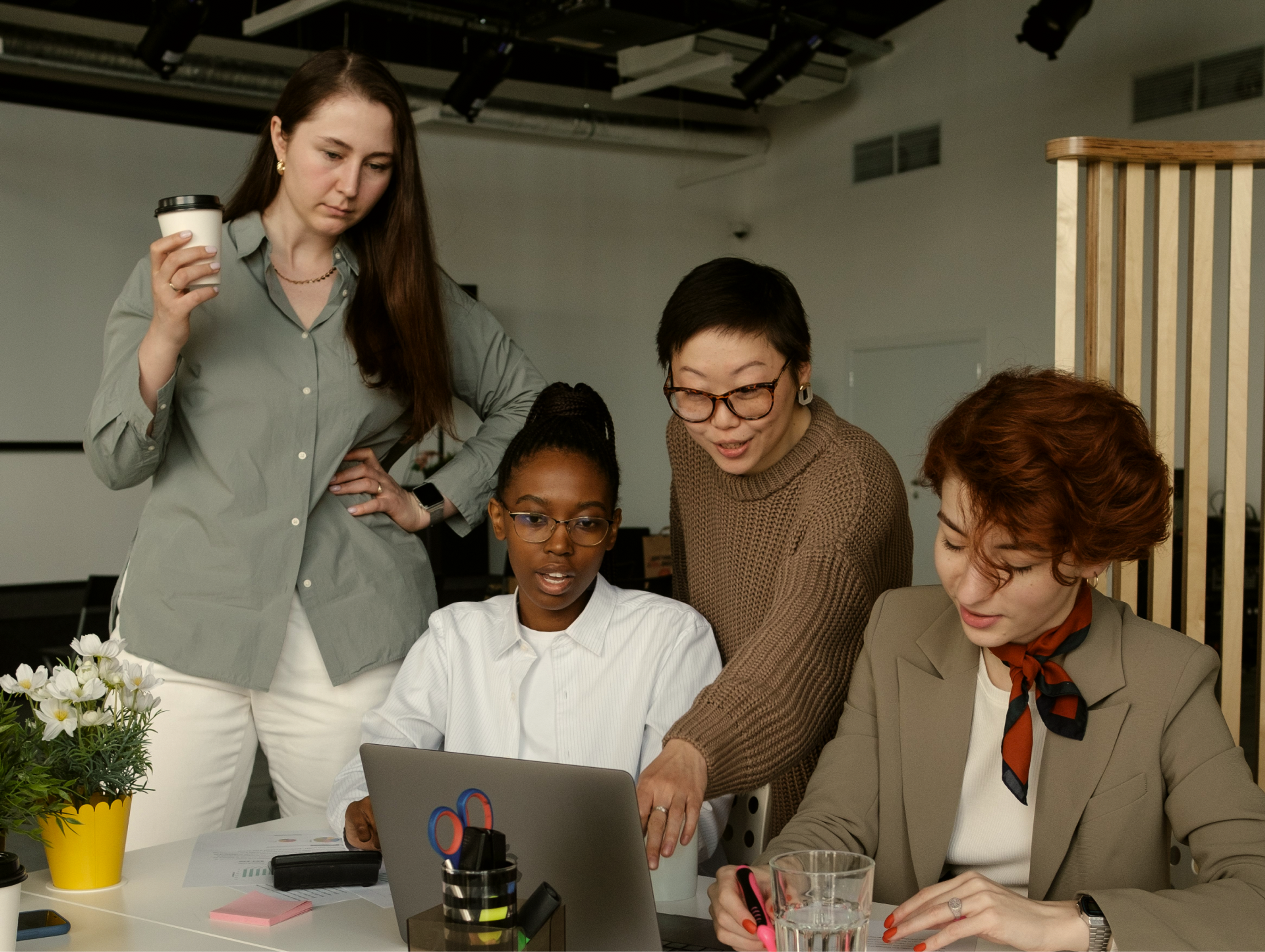 Four women collaborate at a desk with a laptop. One holds a coffee cup, another points at the screen, while the others watch and take notes. Office supplies and a small plant are on the table.