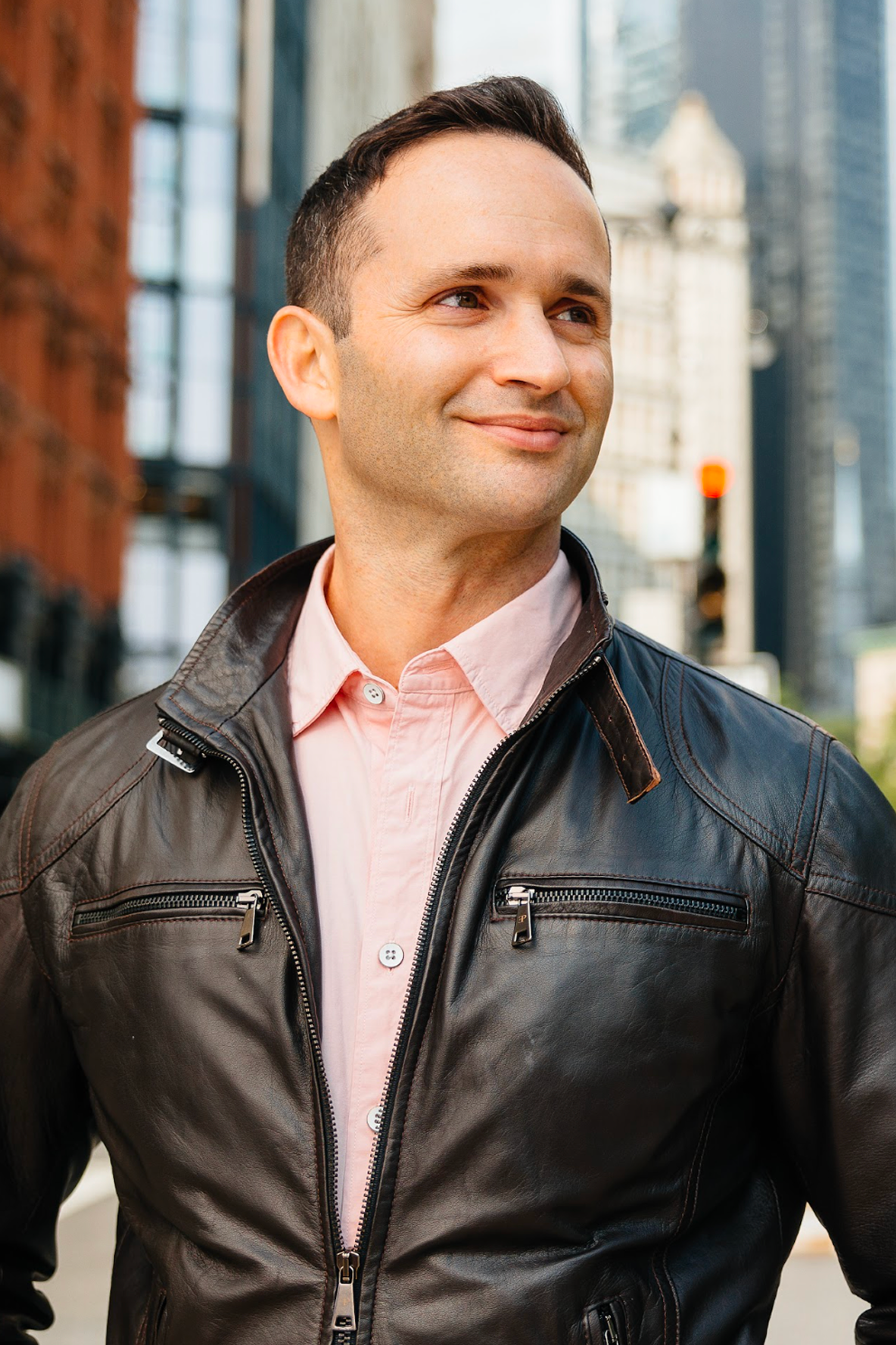 A man wearing a black leather jacket and pink shirt stands outdoors in a city, smiling slightly while looking to the side. Tall buildings and a traffic light are visible in the blurred background.