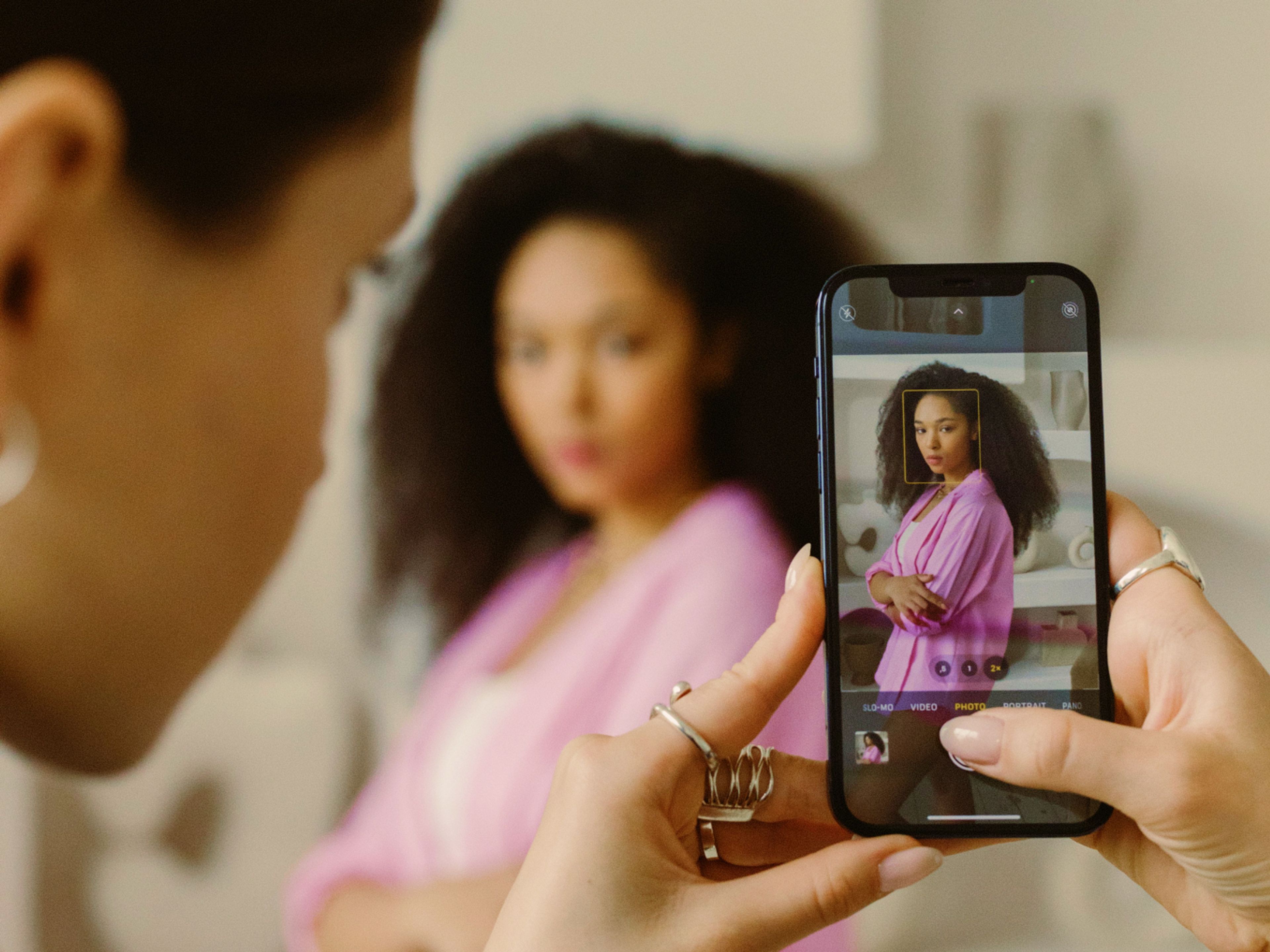 A person holds a phone, taking a photo of a woman with curly hair who is wearing a pink top. The phone screen shows her clearly, while she appears blurred in the background.