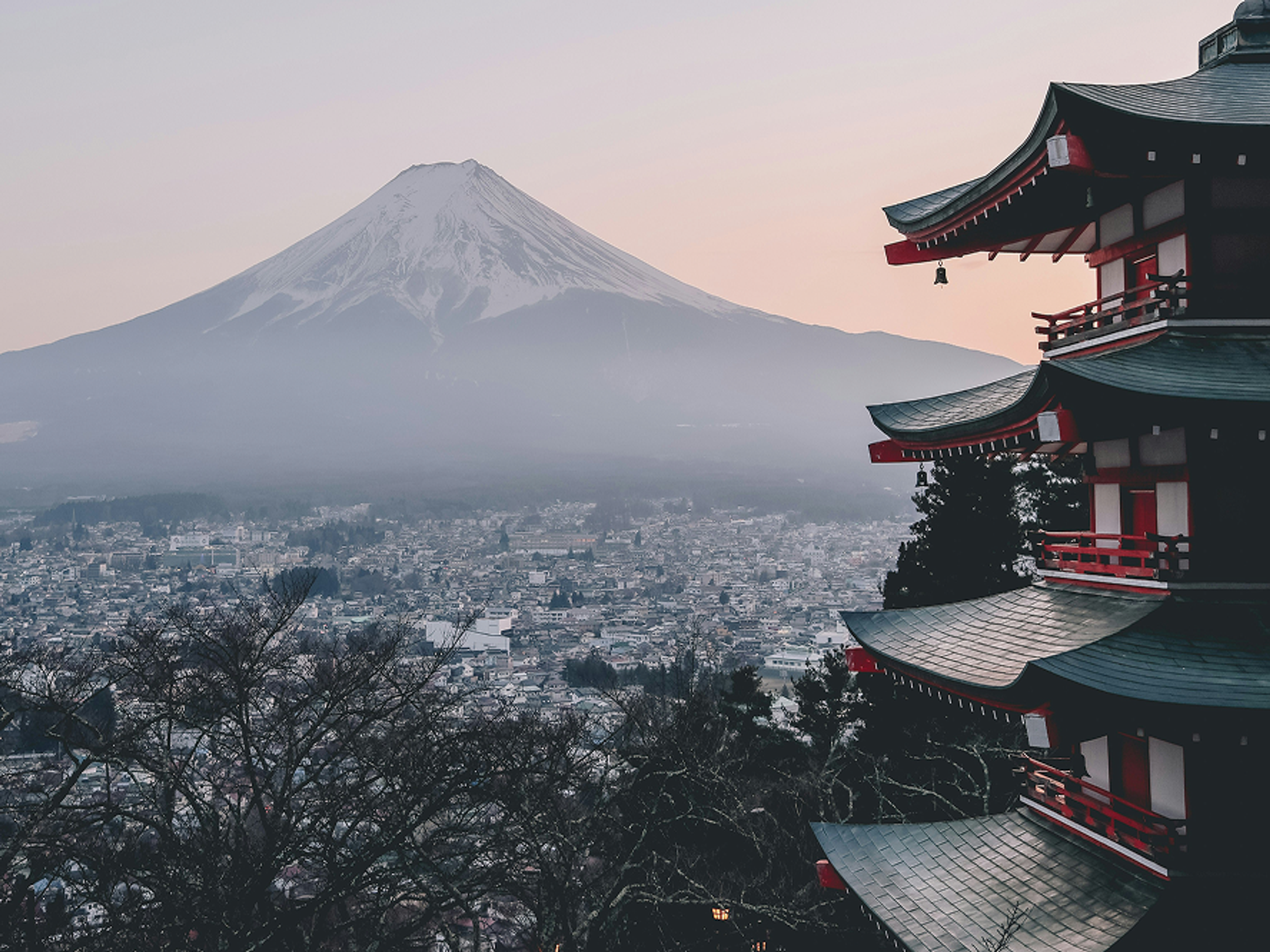 A five-story pagoda in the foreground with Mount Fuji in the background at sunset, overlooking a city partially covered in mist and surrounded by bare trees.