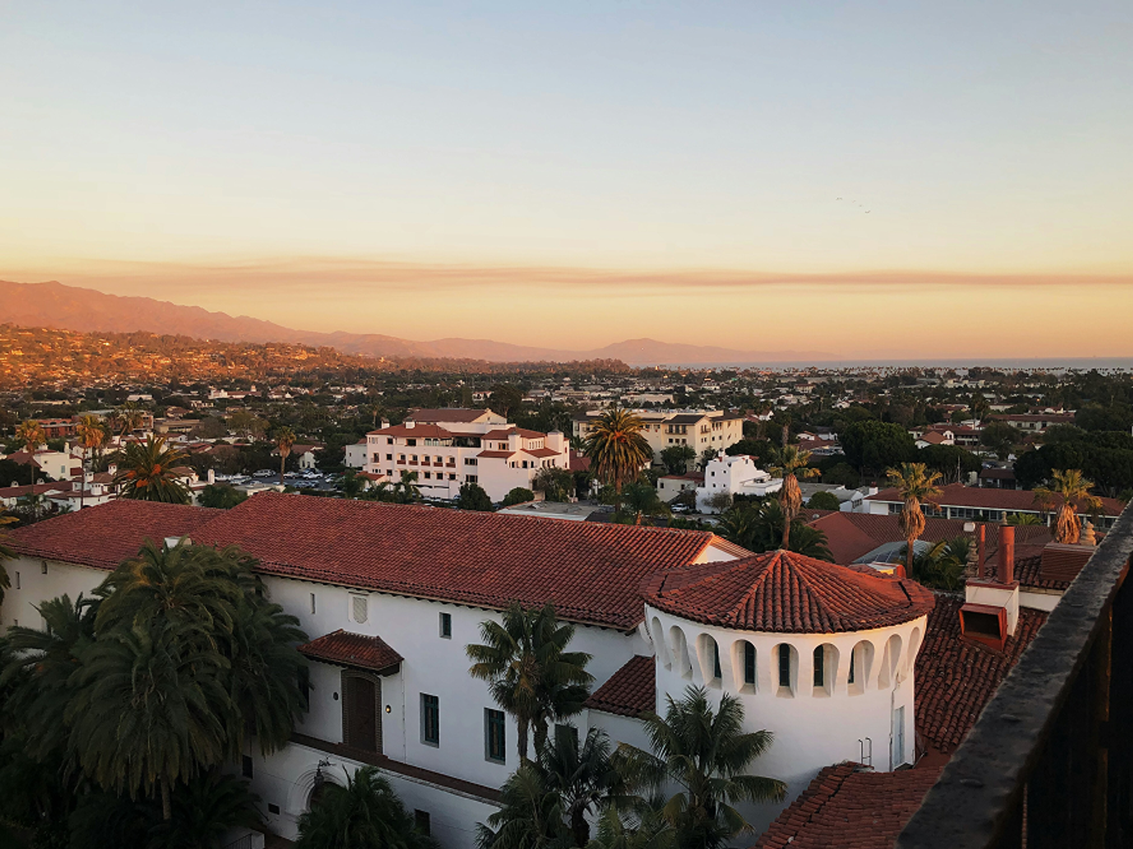 A scenic view of a city at sunset, featuring red-tiled roofs, white buildings, palm trees, and distant mountains under a soft, orange sky.