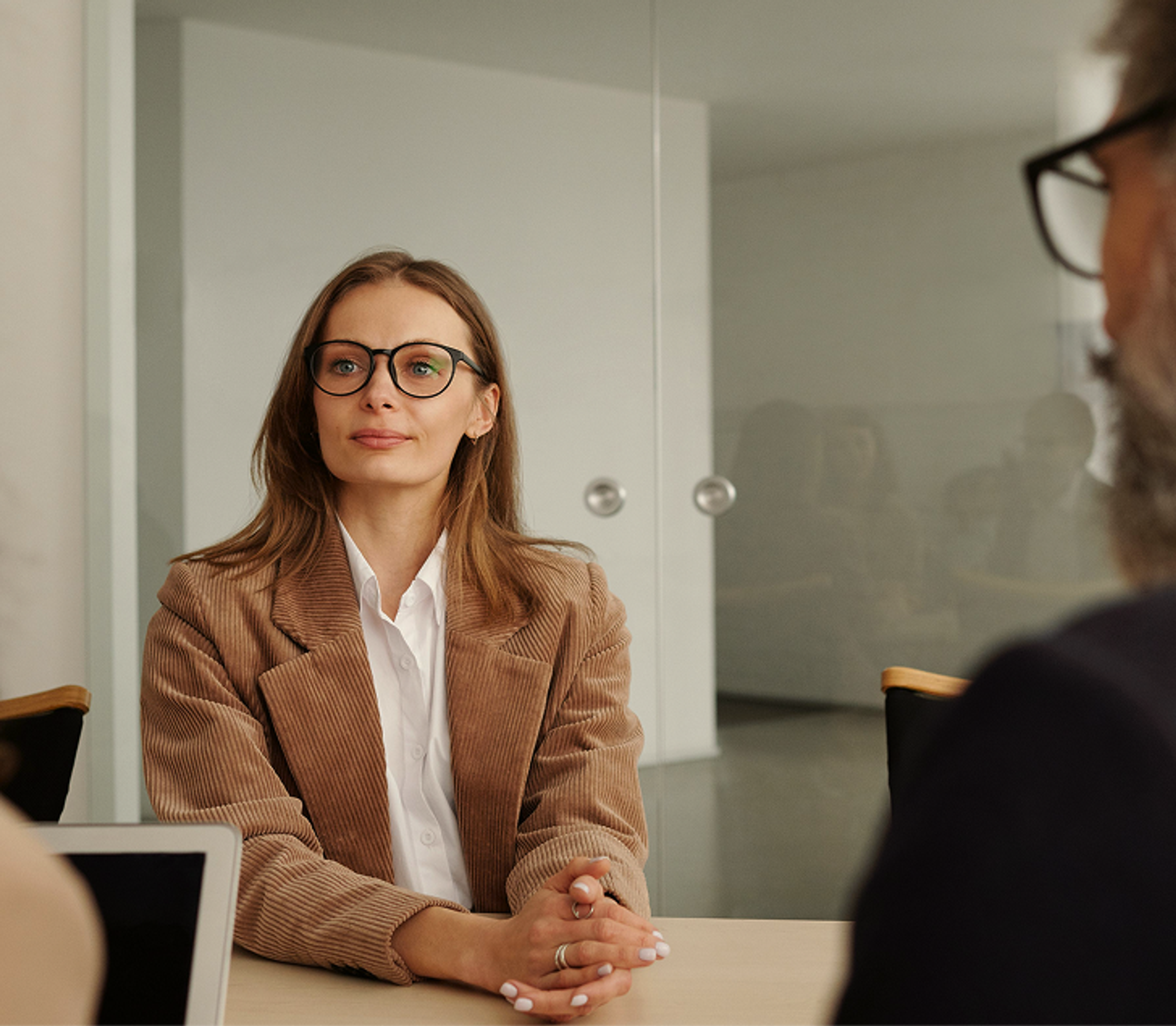 A woman with glasses and a brown blazer sits at a table during a meeting or interview, facing two blurred people across from her in a modern office setting.