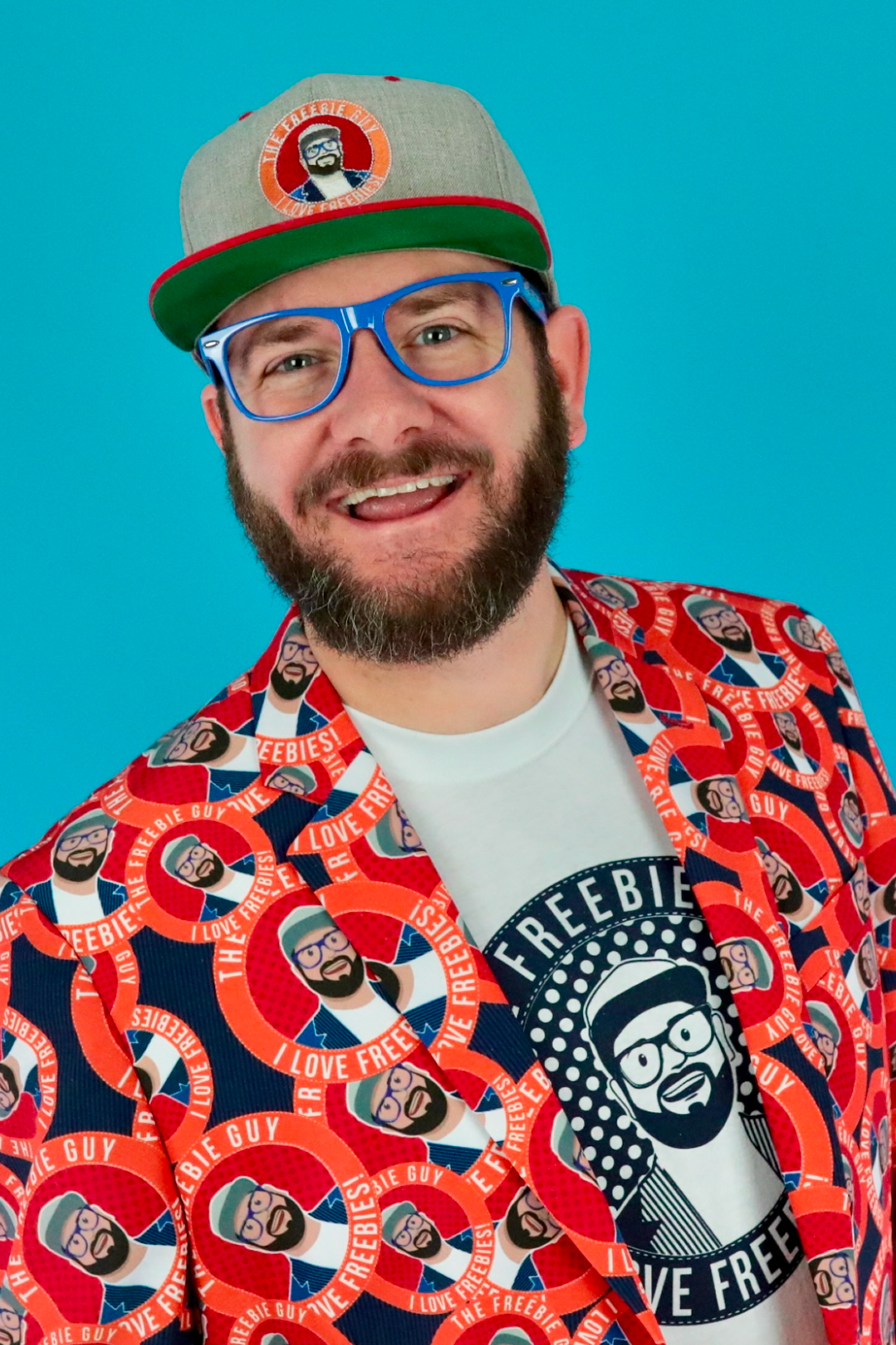 A smiling man wearing blue glasses, a gray cap, and a colorful suit covered with cartoon faces and the words I love freebie guy. His shirt and hat also feature the same cartoon face. Blue background.