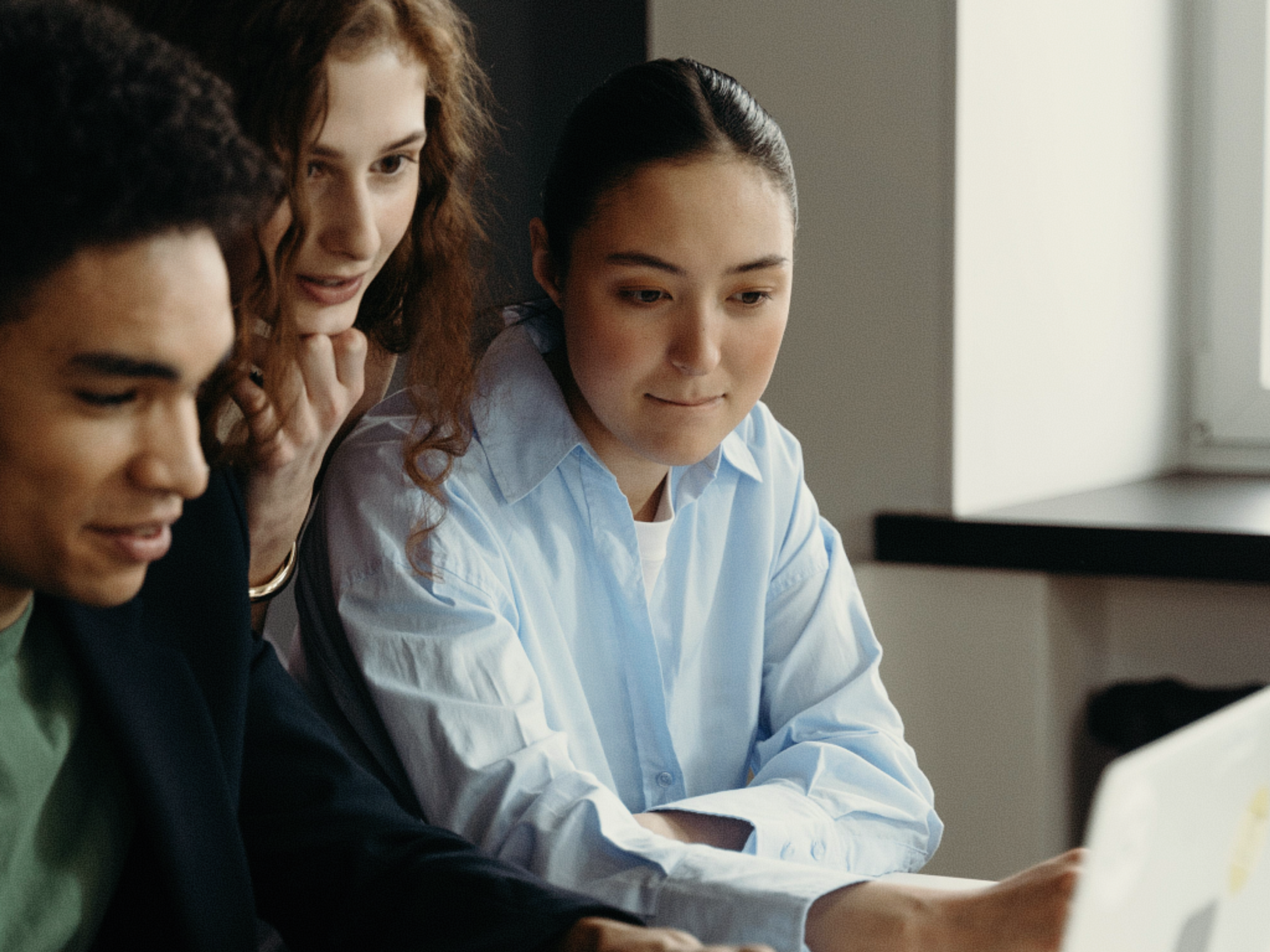 Three people sit closely together, focused on something off-screen, possibly a laptop. Two women and one man appear engaged and thoughtful in a bright room with a window in the background.