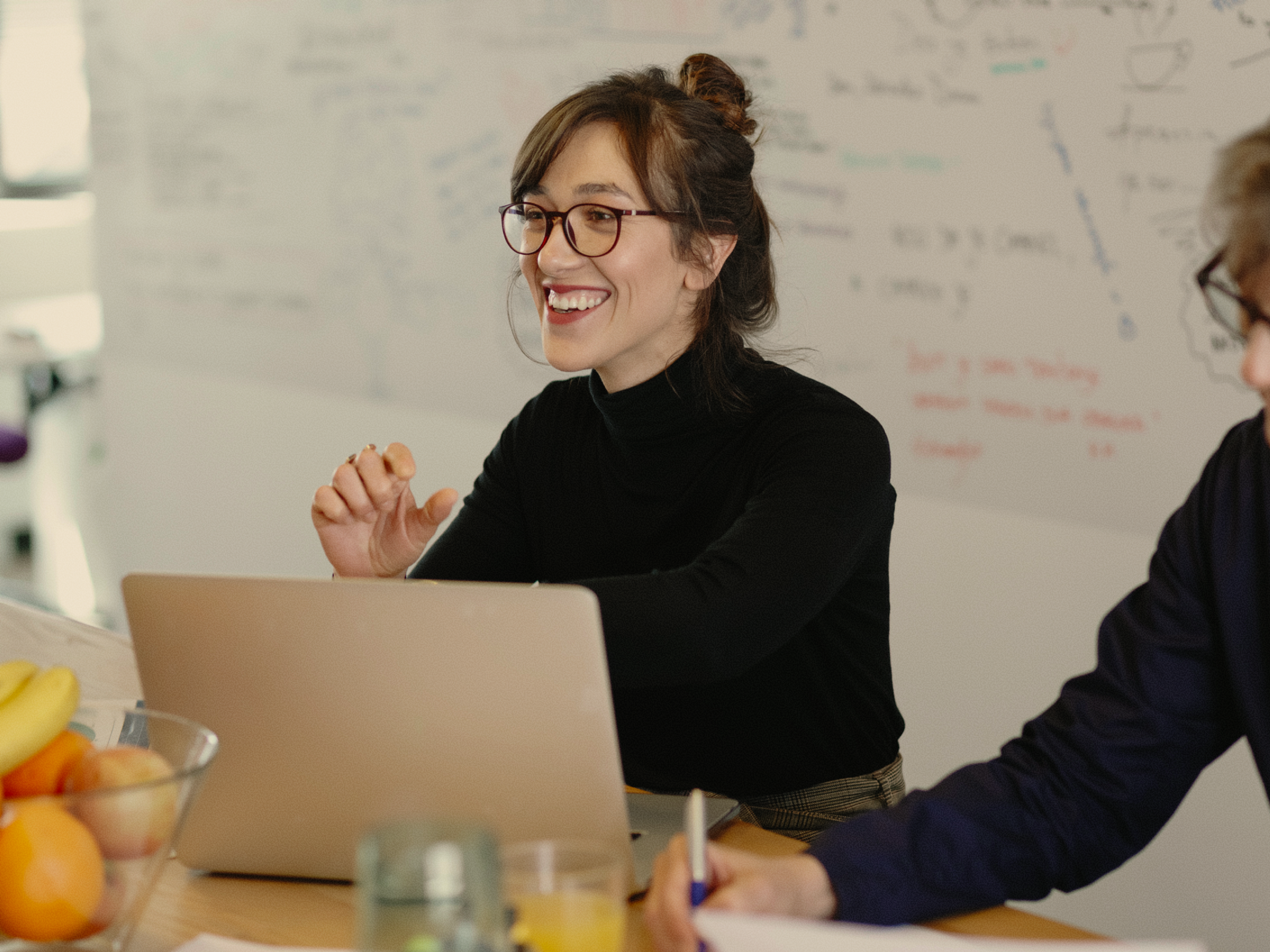 A woman wearing glasses and a black top smiles while sitting at a table with a laptop. There is a bowl of fruit and a whiteboard with notes in the background.