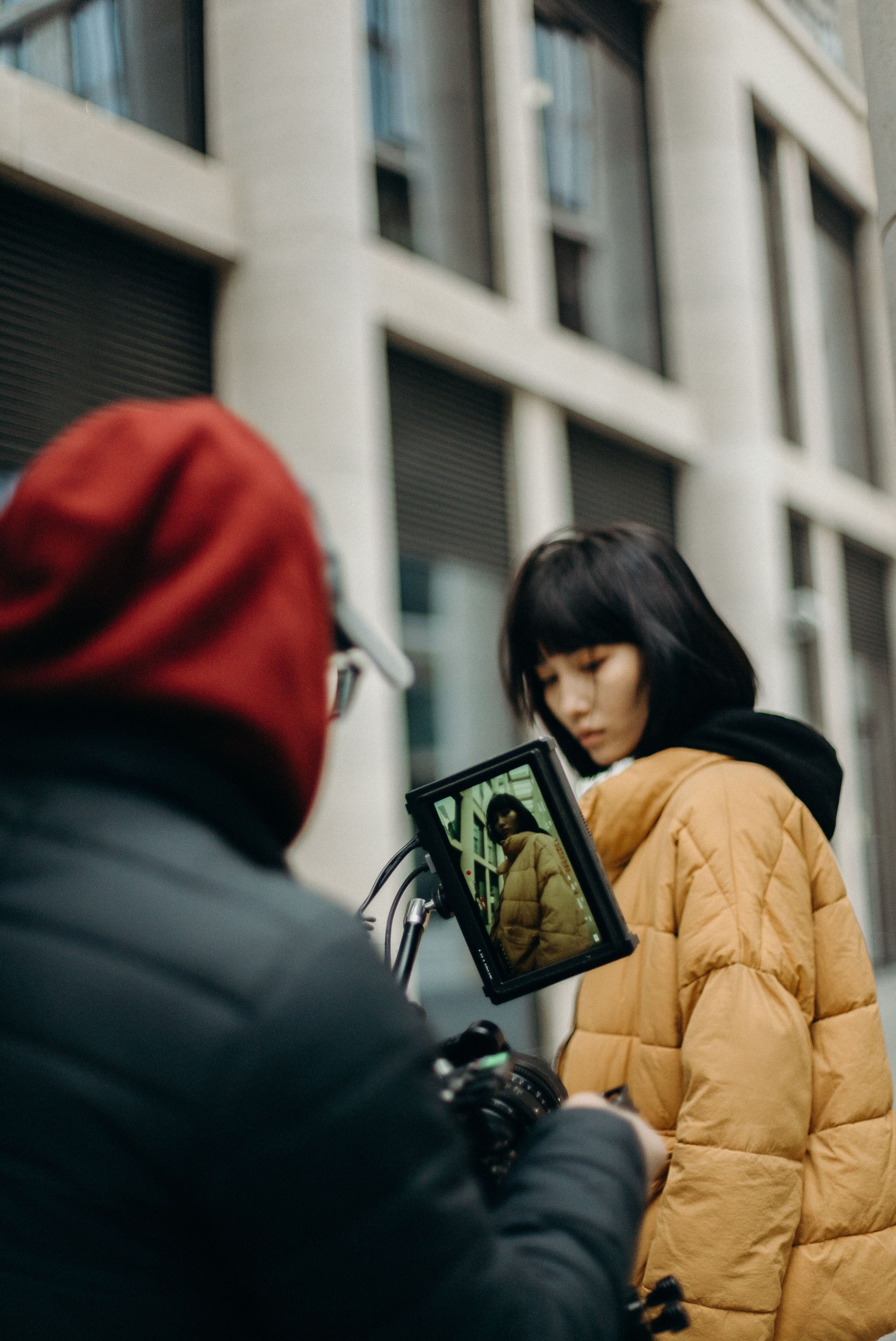 A person in a yellow jacket looks down while being filmed outdoors; the camera operator in a red hood captures their image, which appears on a monitor.