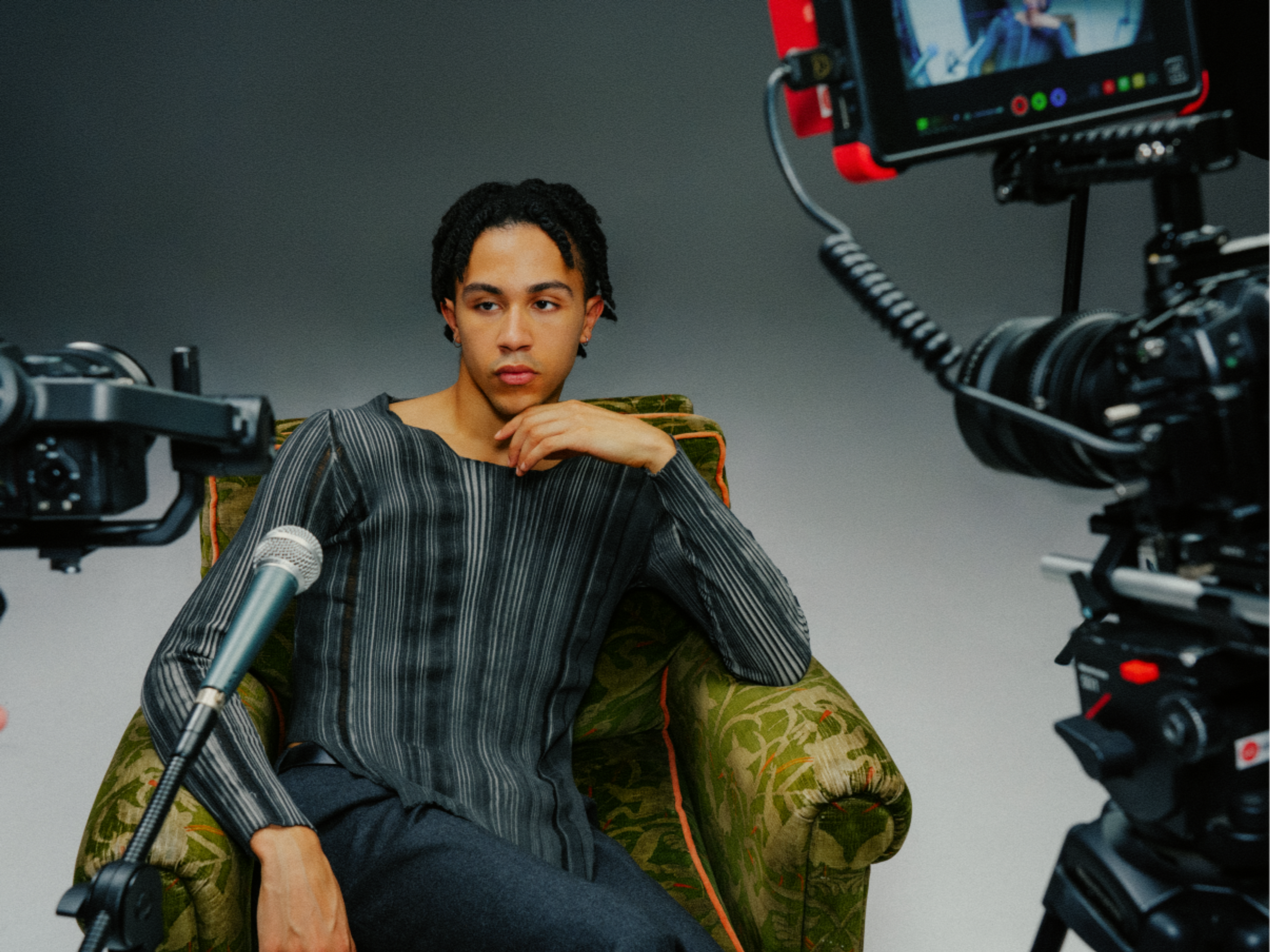A person sits on a green patterned armchair, posing thoughtfully for an interview while surrounded by cameras and a microphone in a studio setting.