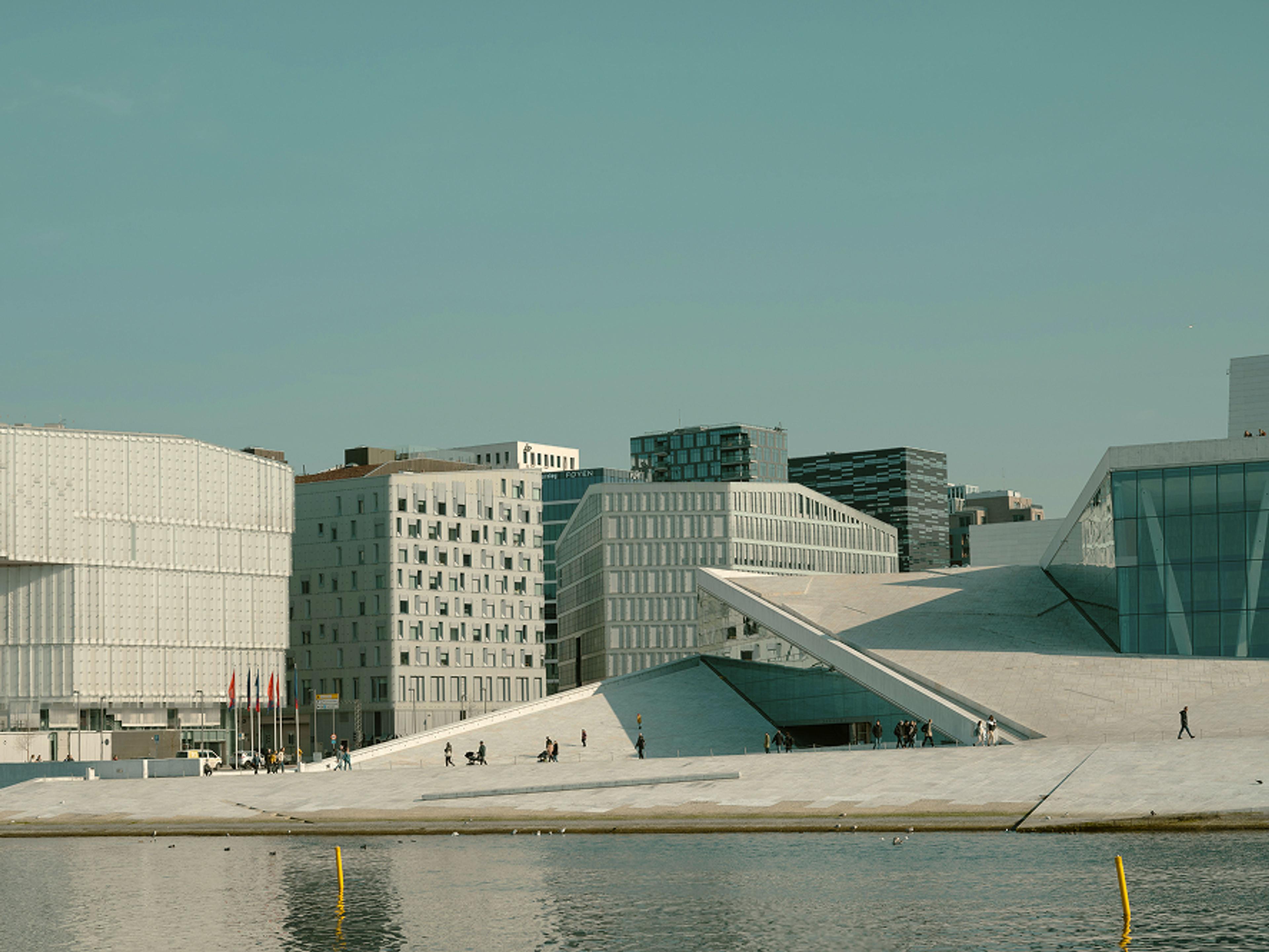 Modern waterfront buildings with geometric shapes and sloped surfaces, people walking on wide white steps, and calm water in the foreground under a clear sky.