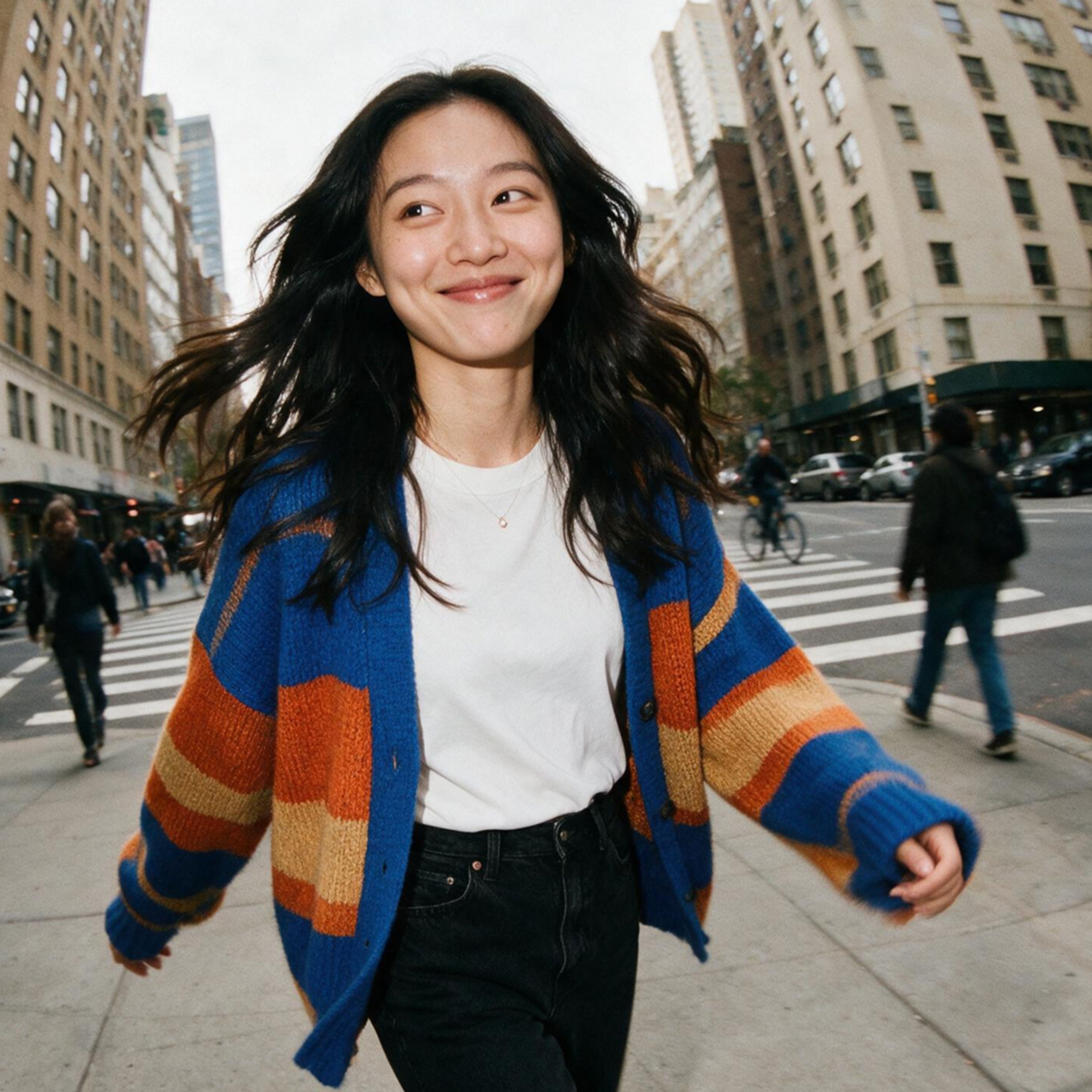 A woman with long dark hair, wearing a striped cardigan and white t-shirt, smiles while walking across a city street lined with tall buildings and people.