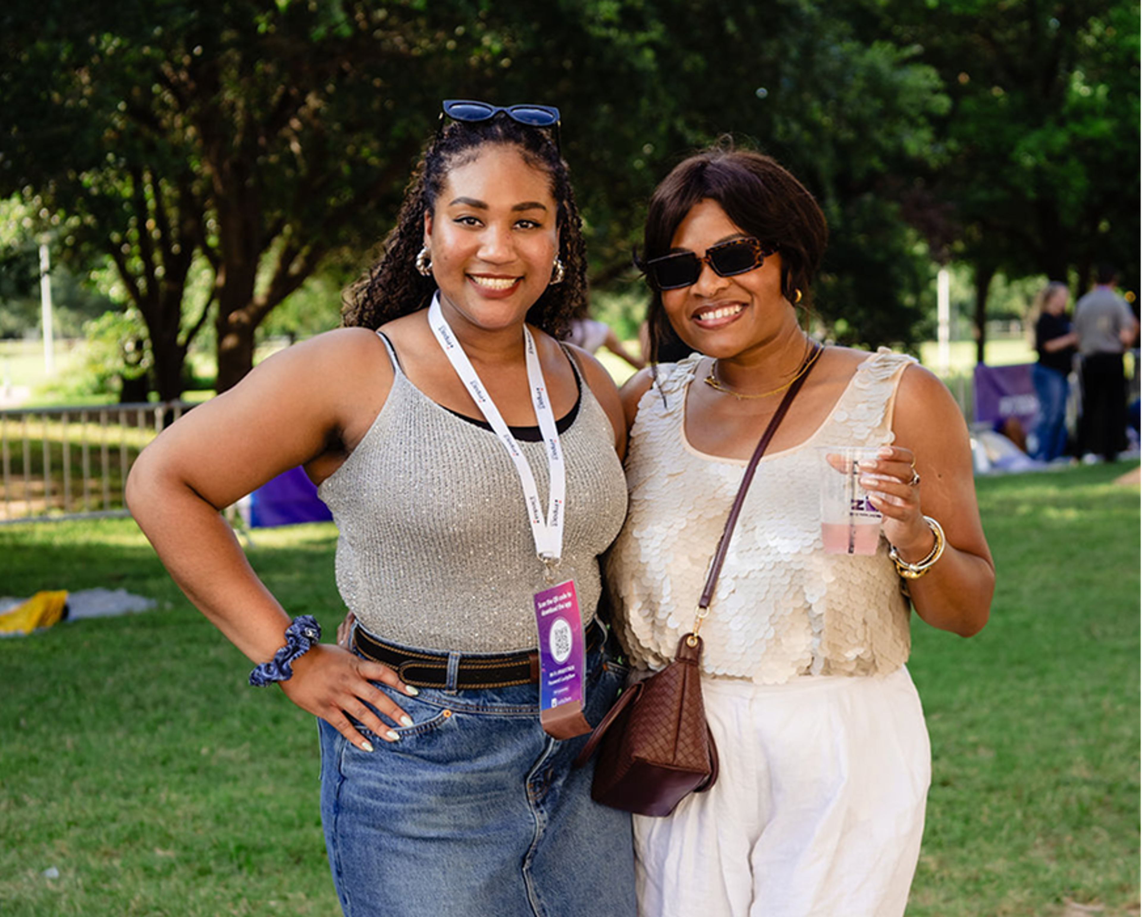Two women smiling outdoors, wearing summer outfits and sunglasses. One has a lanyard and event badge, the other holds a drink. Green grass and trees are in the background.