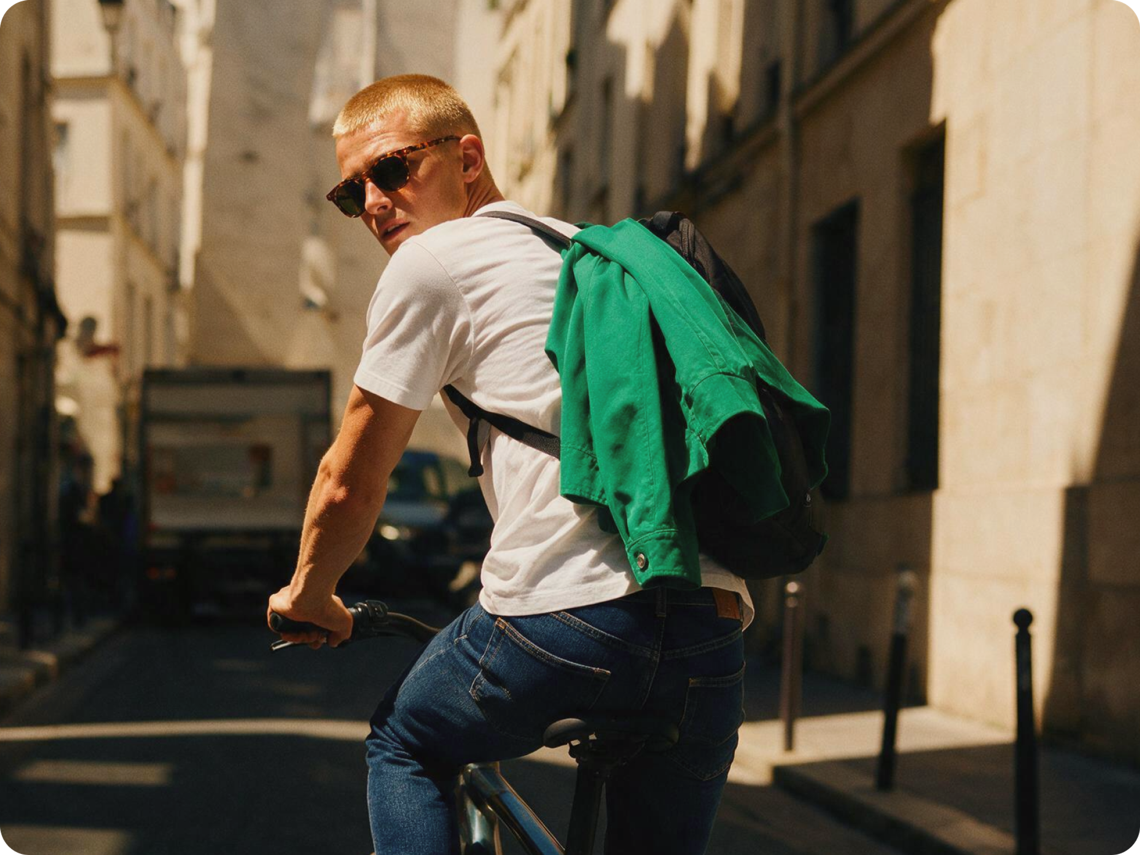 A person wearing sunglasses, a white t-shirt, and jeans rides a bicycle down a city street with a green jacket draped over their shoulders, looking back over their shoulder.