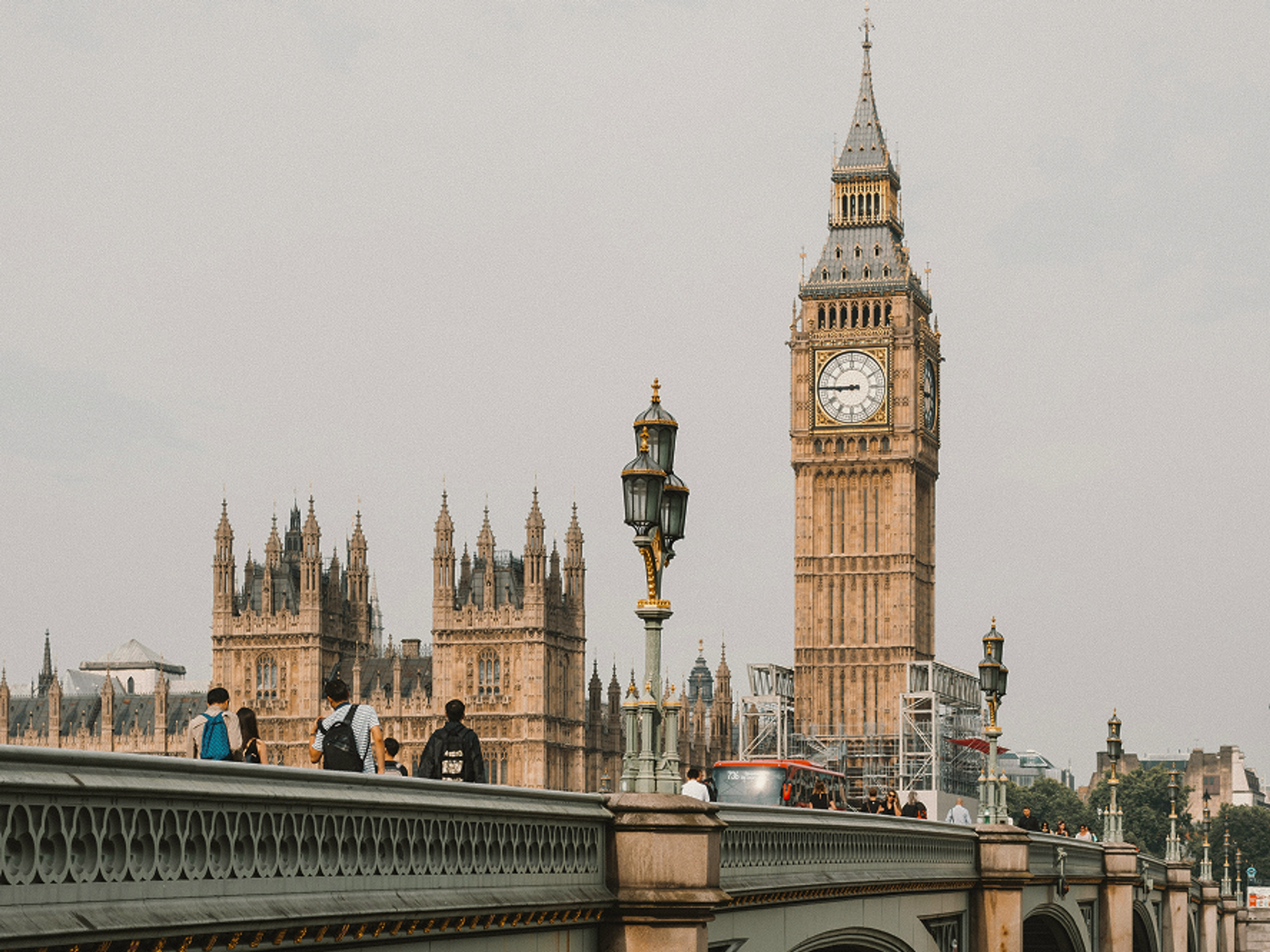 People walk across Westminster Bridge in London. The Houses of Parliament and the Elizabeth Tower, commonly known as Big Ben, are visible in the background on a cloudy day.