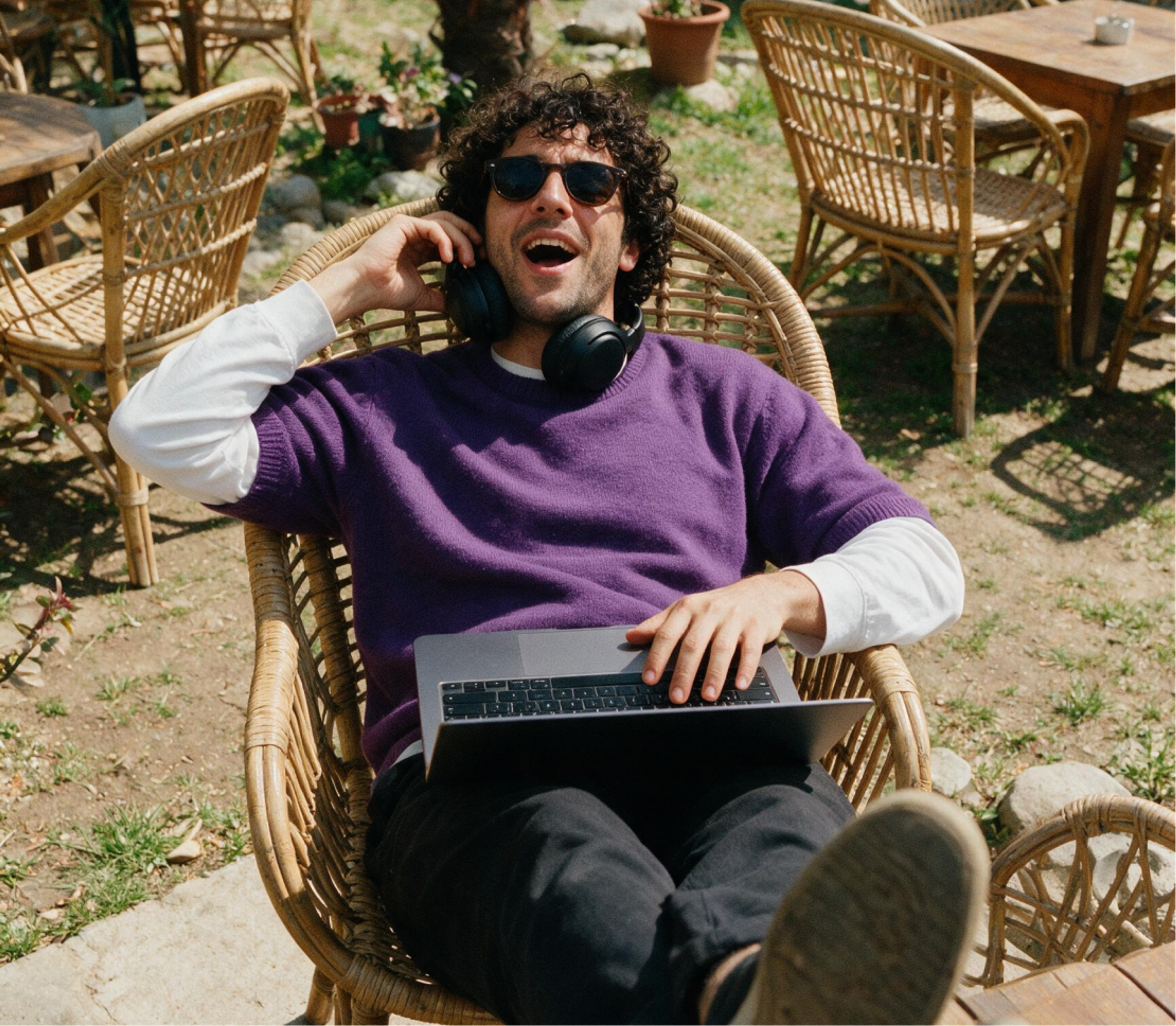 A person wearing sunglasses and headphones relaxes in a wicker chair outdoors, smiling while using a laptop on their lap. There are empty tables and plants in the background.