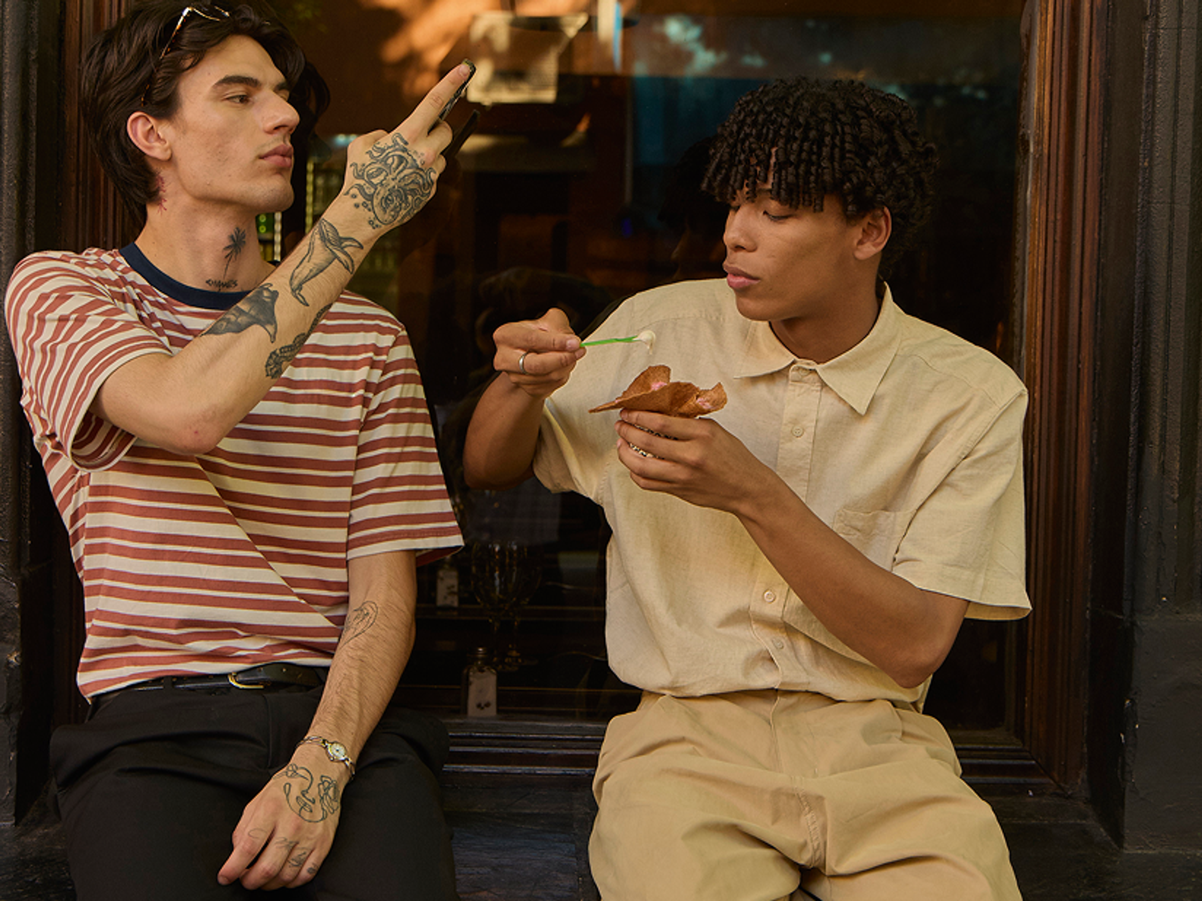 Two young men sit outside. One in a striped shirt gestures with his hand, while the other in a beige shirt eats dessert from a bowl with a spoon. They appear relaxed and are interacting with each other.