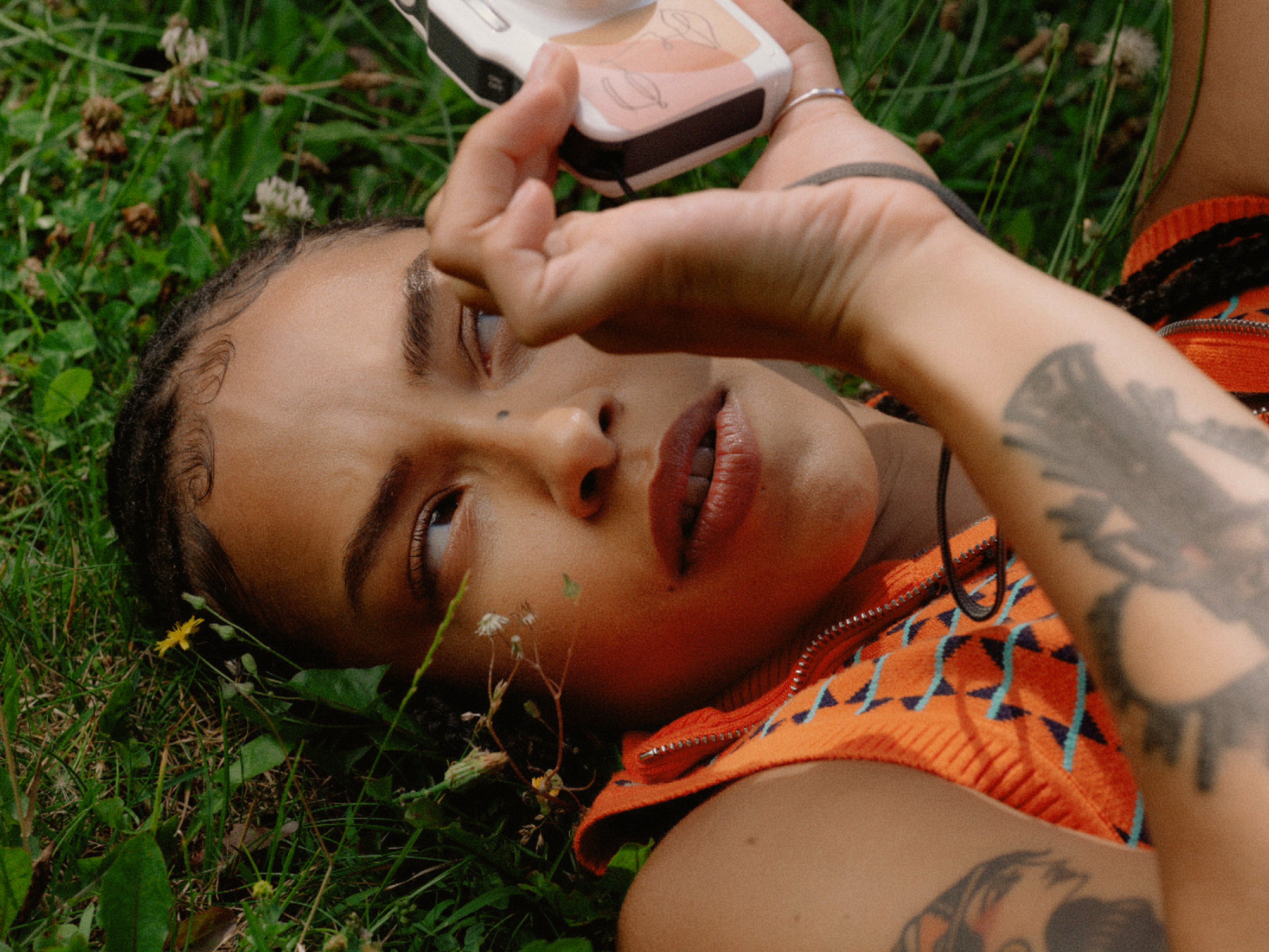 A person with braids and visible tattoos lies on grass, looking intently at a camera or phone held above their face, wearing an orange patterned top.
