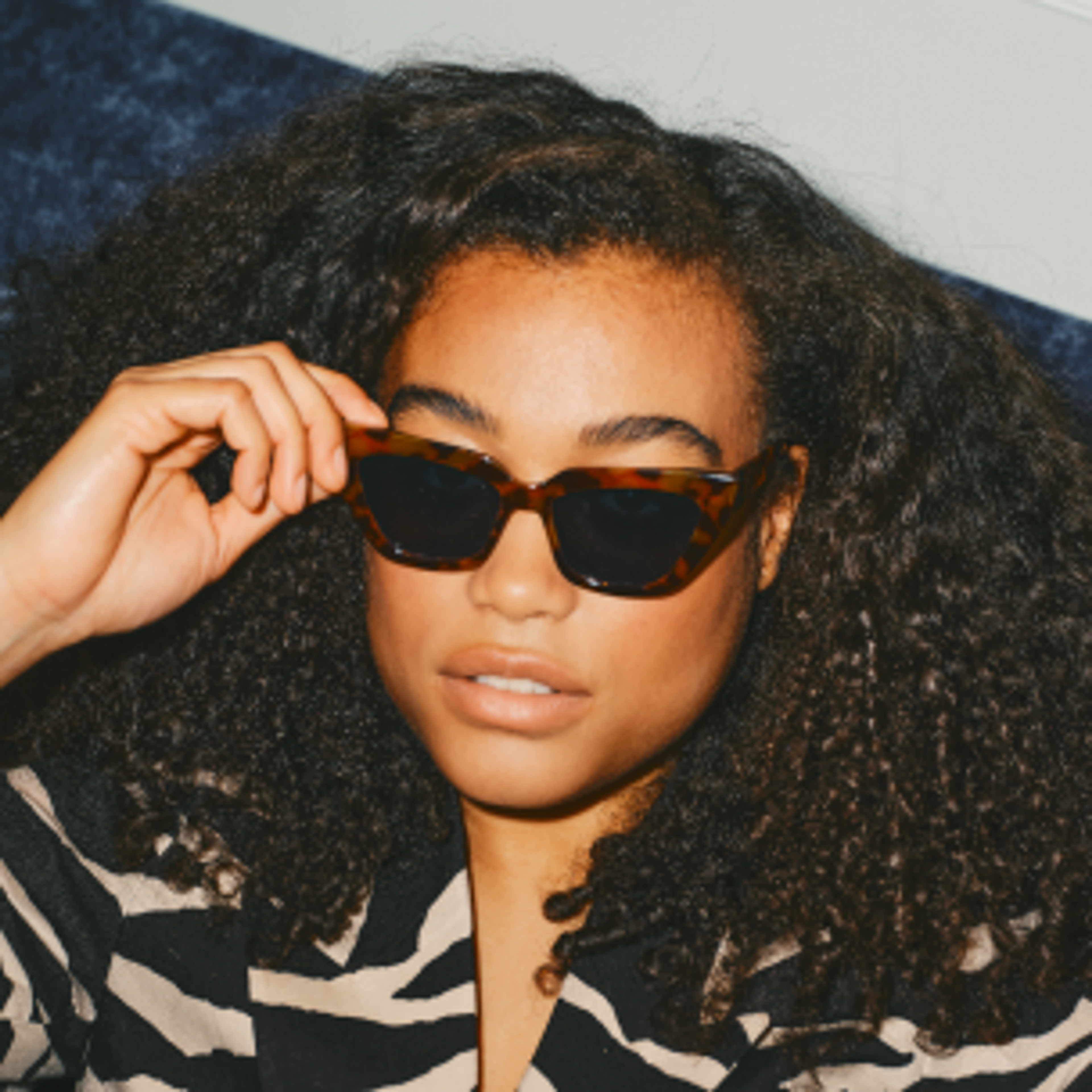 A person with curly hair wearing tortoiseshell sunglasses and a zebra-patterned top looks at the camera, raising one hand to adjust their glasses.