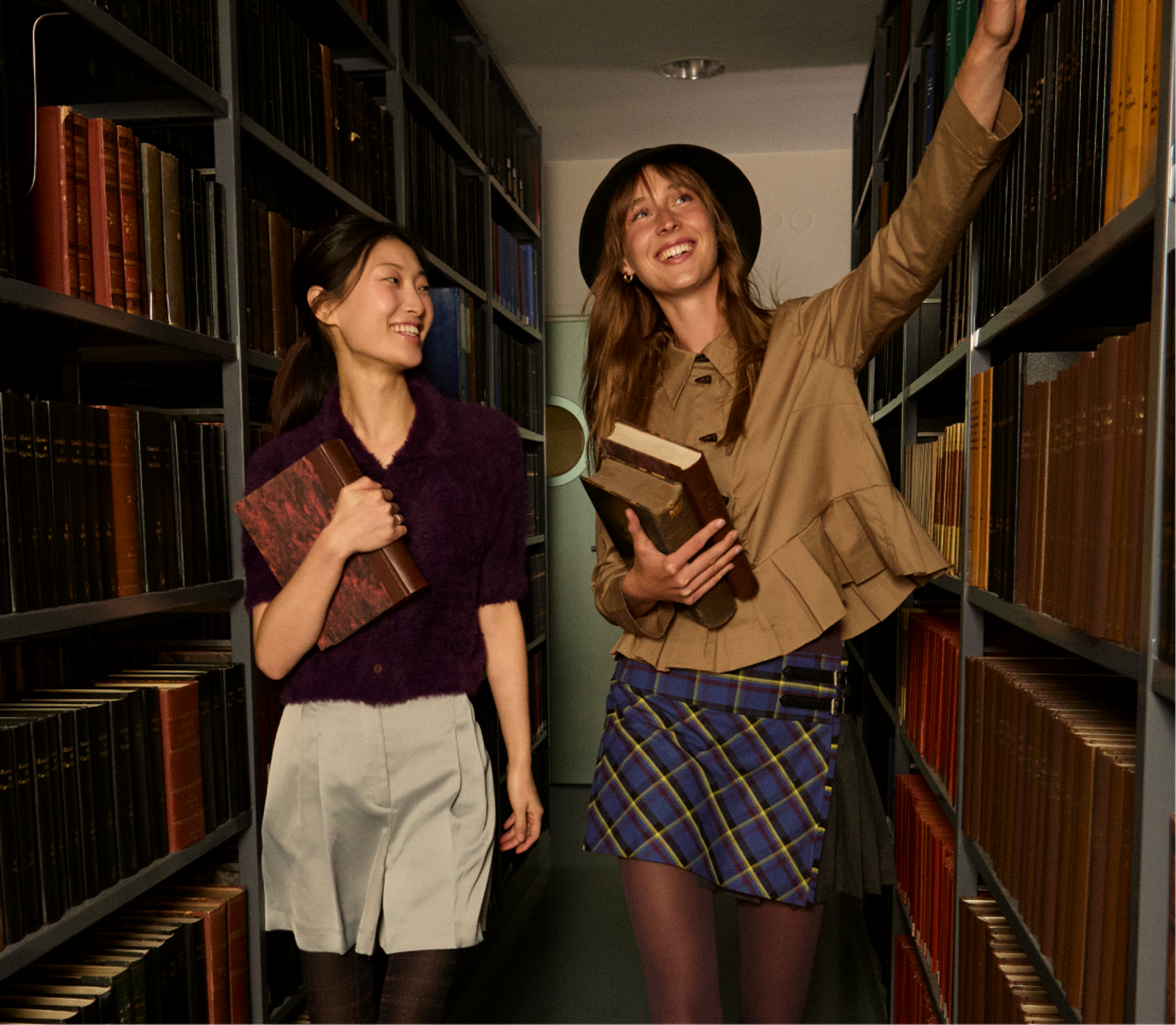 Two young women stand in a library aisle, smiling. One holds books and points at a high shelf, while the other looks at her, also holding a book. Bookshelves filled with books line both sides of the narrow aisle.