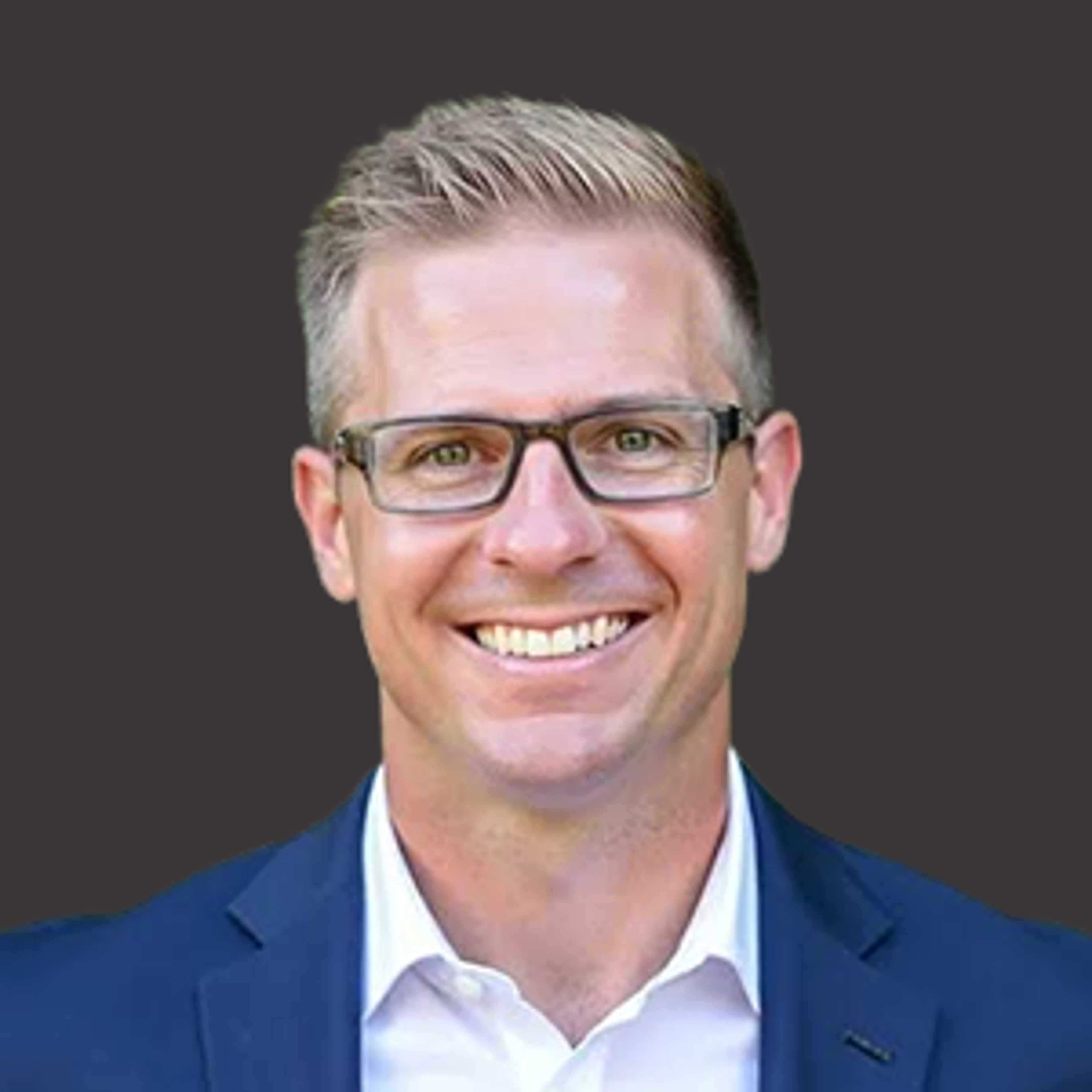 Smiling man with short, light hair and glasses, wearing a navy blue suit jacket and white shirt, against a plain dark background.