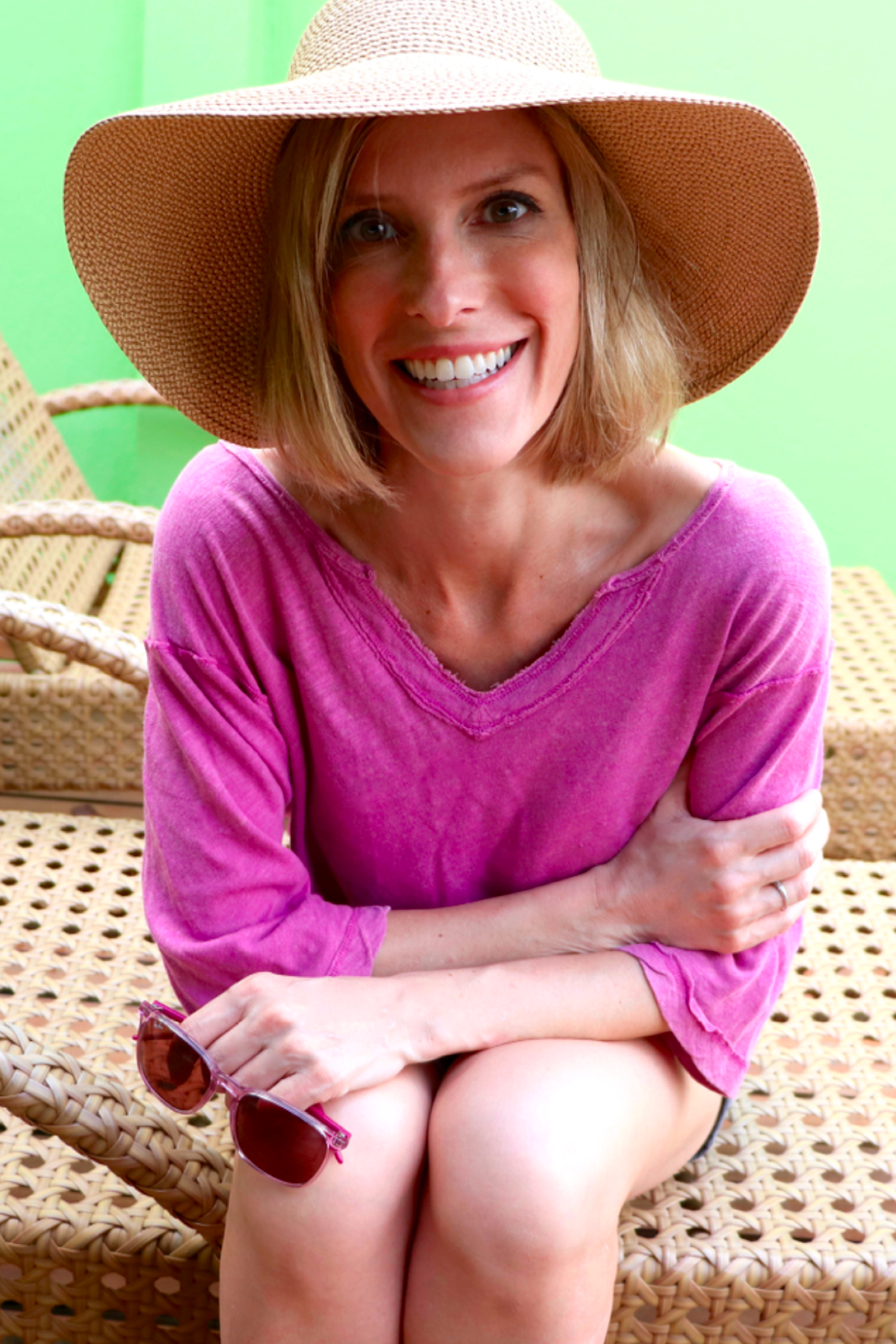 A smiling woman in a wide-brimmed hat and pink top sits on a wicker chair, holding sunglasses in one hand, with a bright green wall behind her.