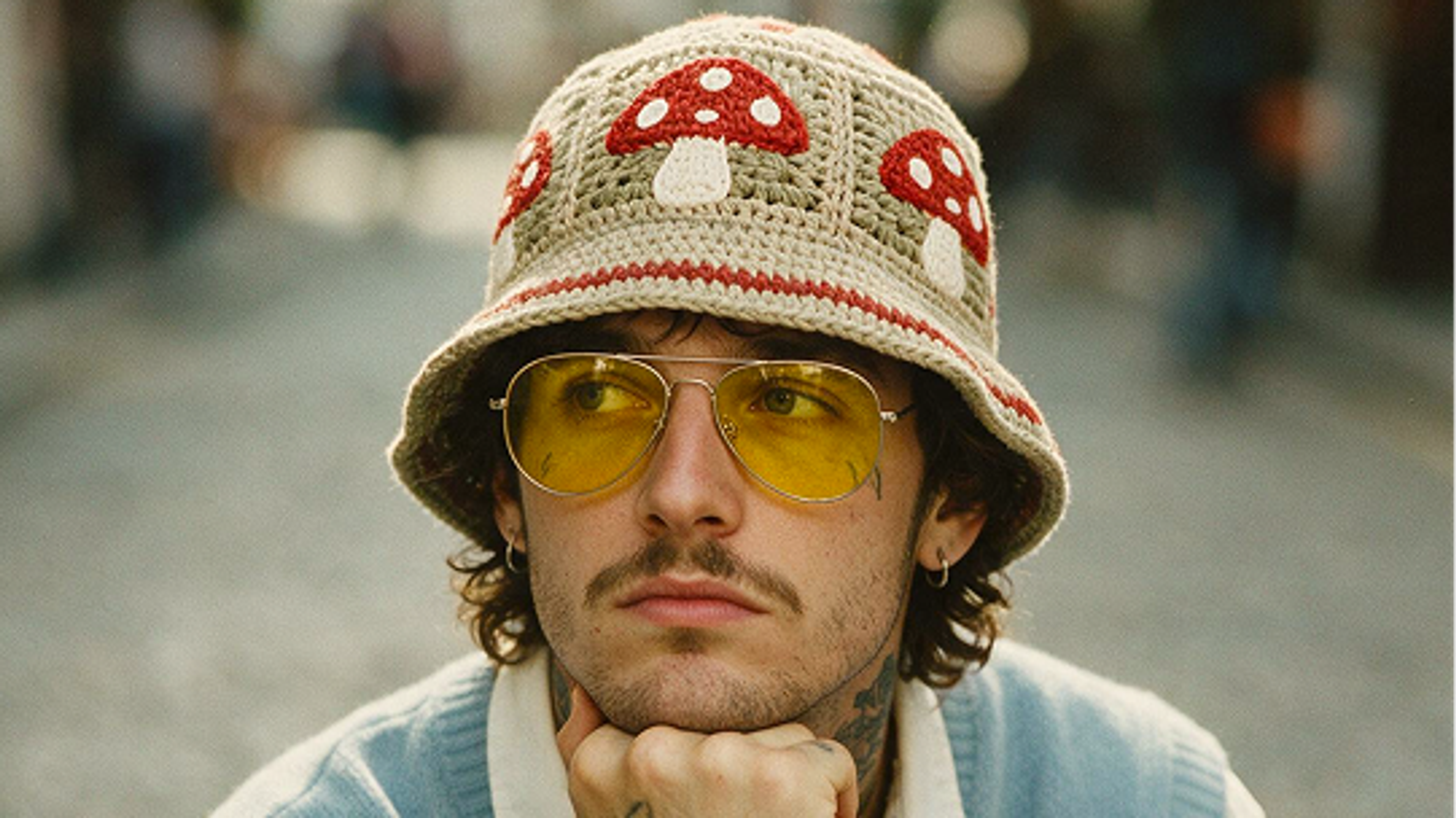 A person with wavy hair, wearing yellow-tinted glasses and a crocheted bucket hat with red and white mushroom designs, rests their chin on their hand while looking thoughtfully into the distance.