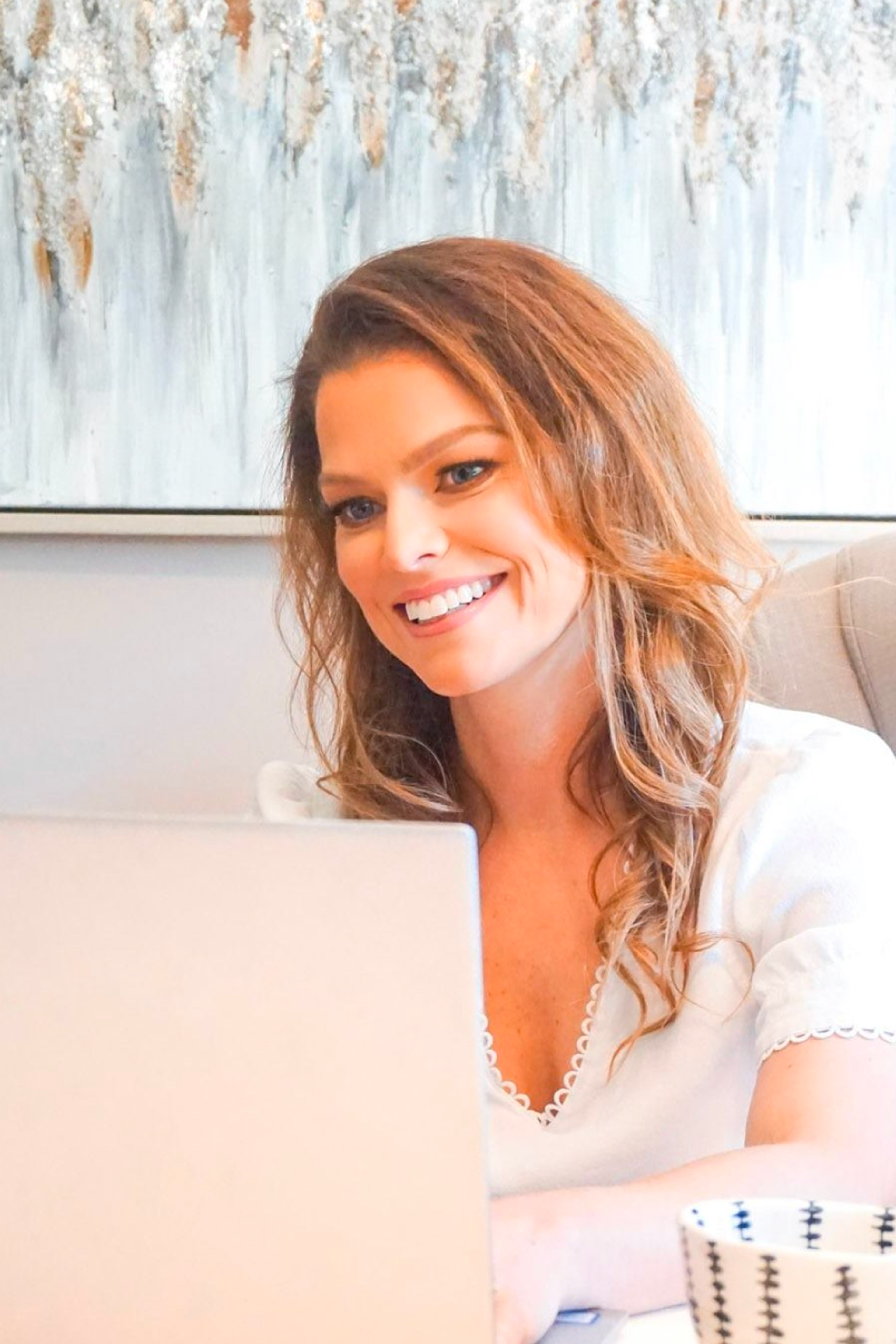 A woman with wavy brown hair and a white top smiles while working at a laptop, with a patterned mug and abstract art in the background.