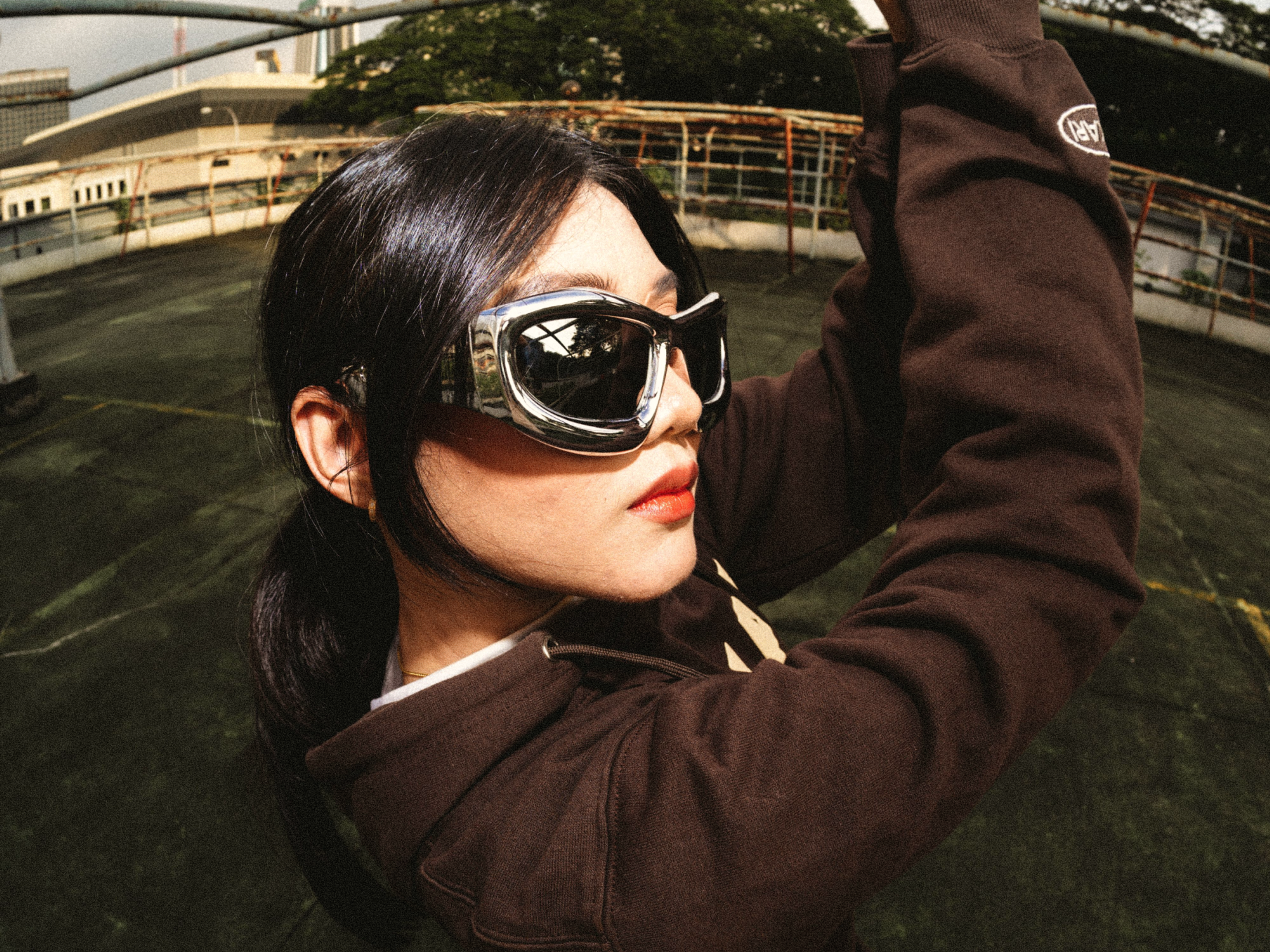 A person with dark hair and red lipstick wears large, reflective sunglasses and a brown jacket, posing outdoors on a rooftop with green trees and buildings in the background.