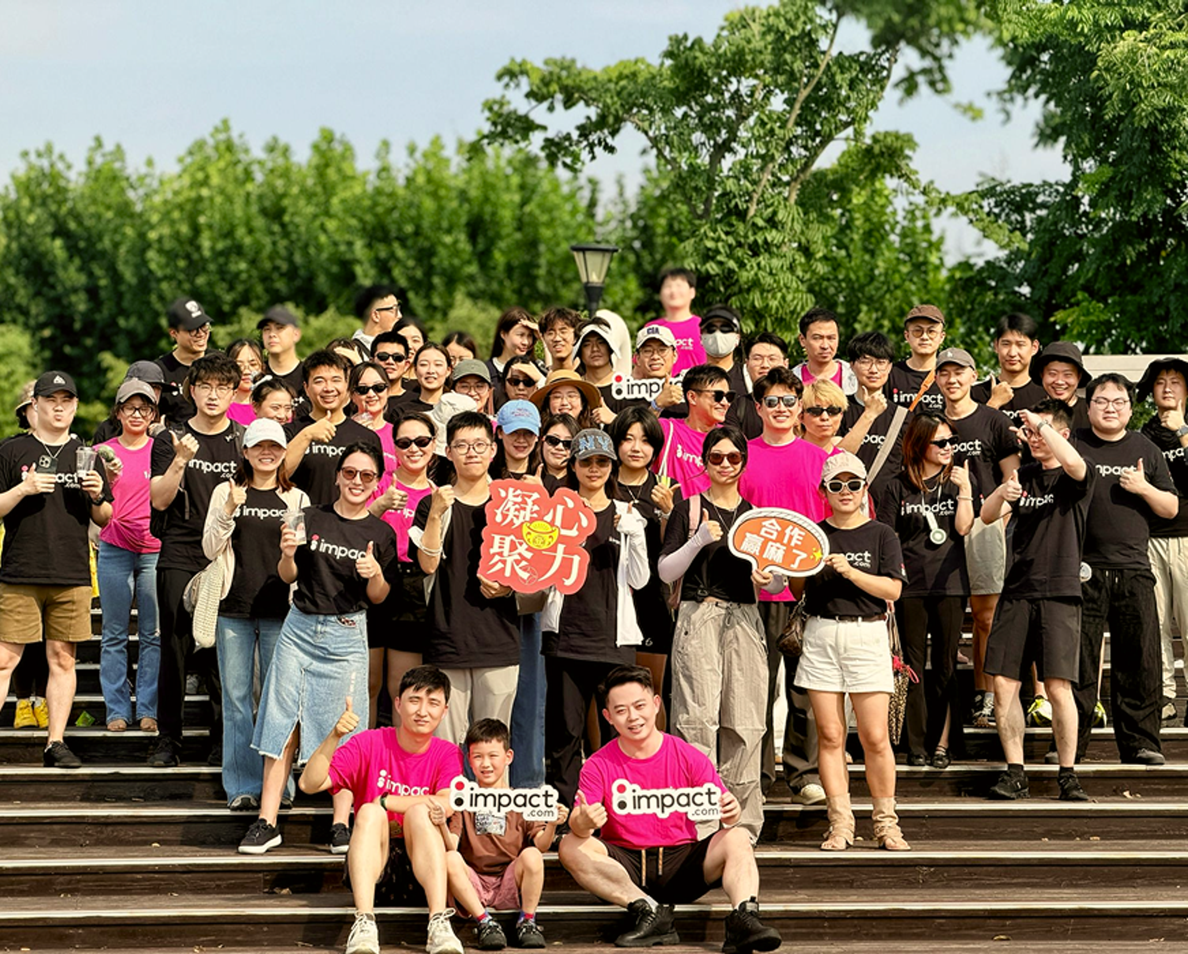A large group of people pose outdoors on wooden steps, wearing black or bright pink impact shirts. Some hold colorful signs with Chinese characters. Trees and a blue sky are in the background.