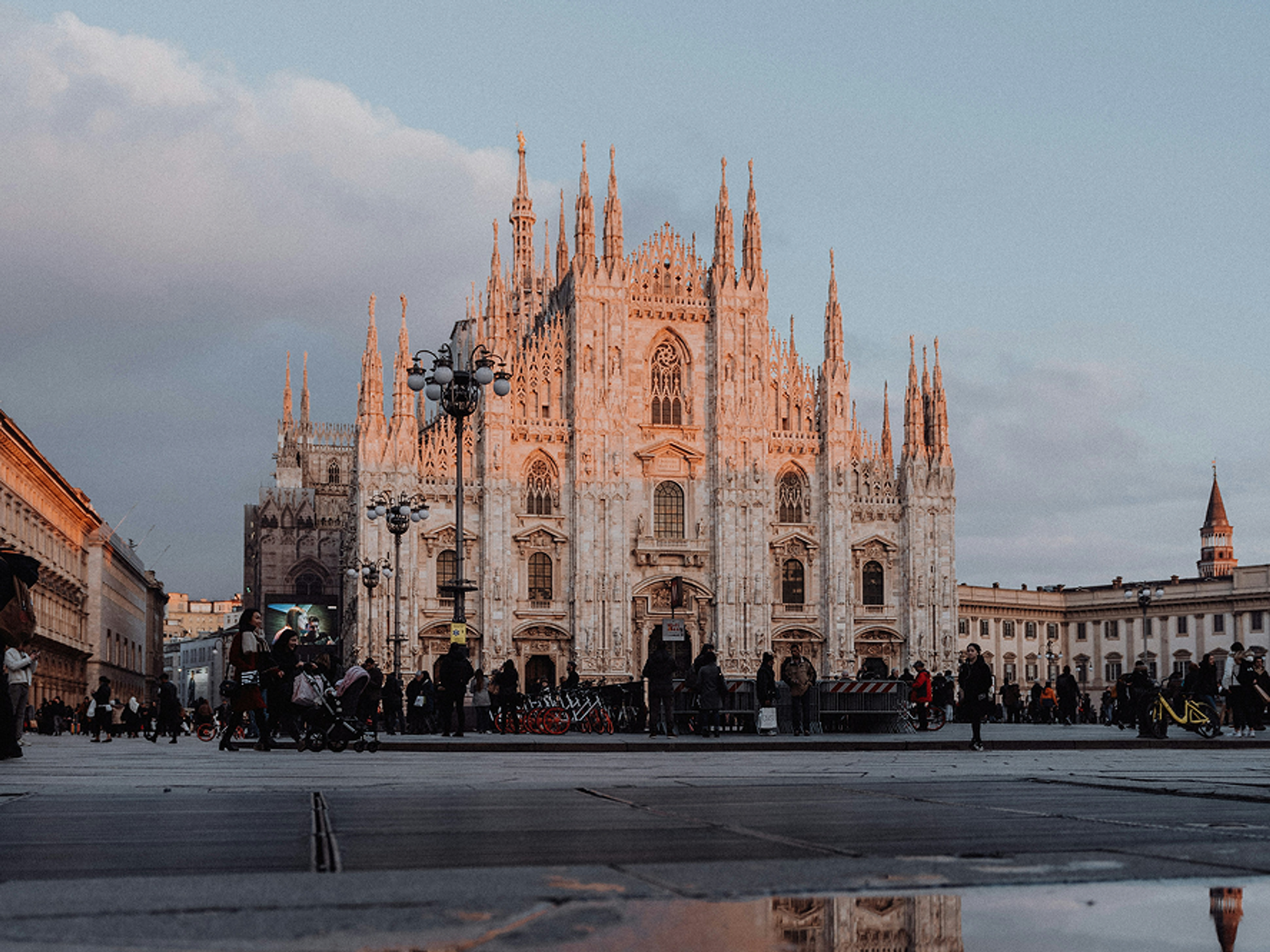 The Milan Cathedral stands in a spacious plaza at sunset, its ornate Gothic facade lit by golden light. People walk and gather in the square, with street lamps and surrounding buildings visible.