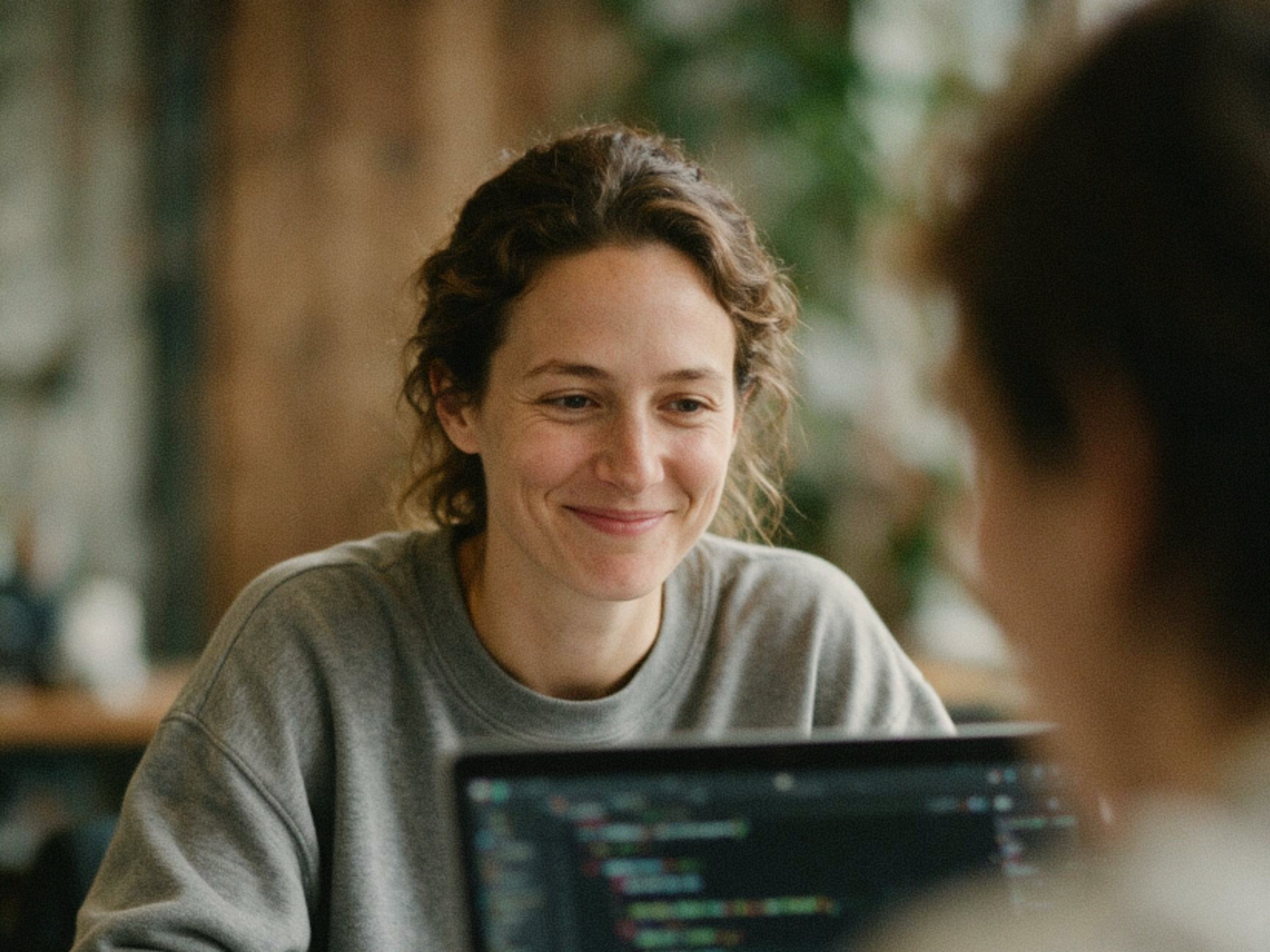 A person with curly hair, wearing a gray sweater, smiles while looking at someone across a table. A blurred laptop screen with code is visible in the foreground. The background is softly out of focus.