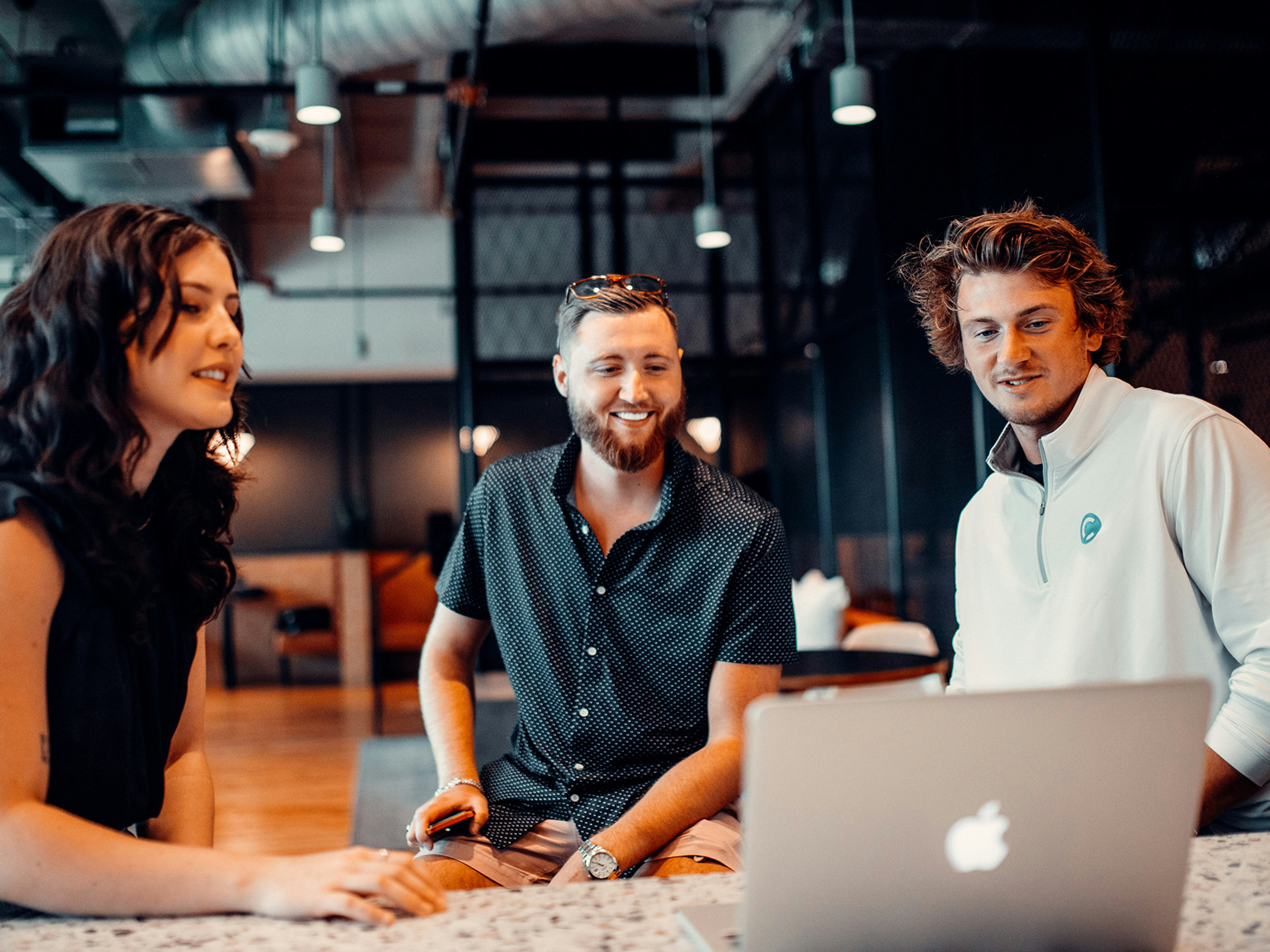 Three young adults sit at a table, smiling and looking at a laptop screen in a modern, industrial-style office space.