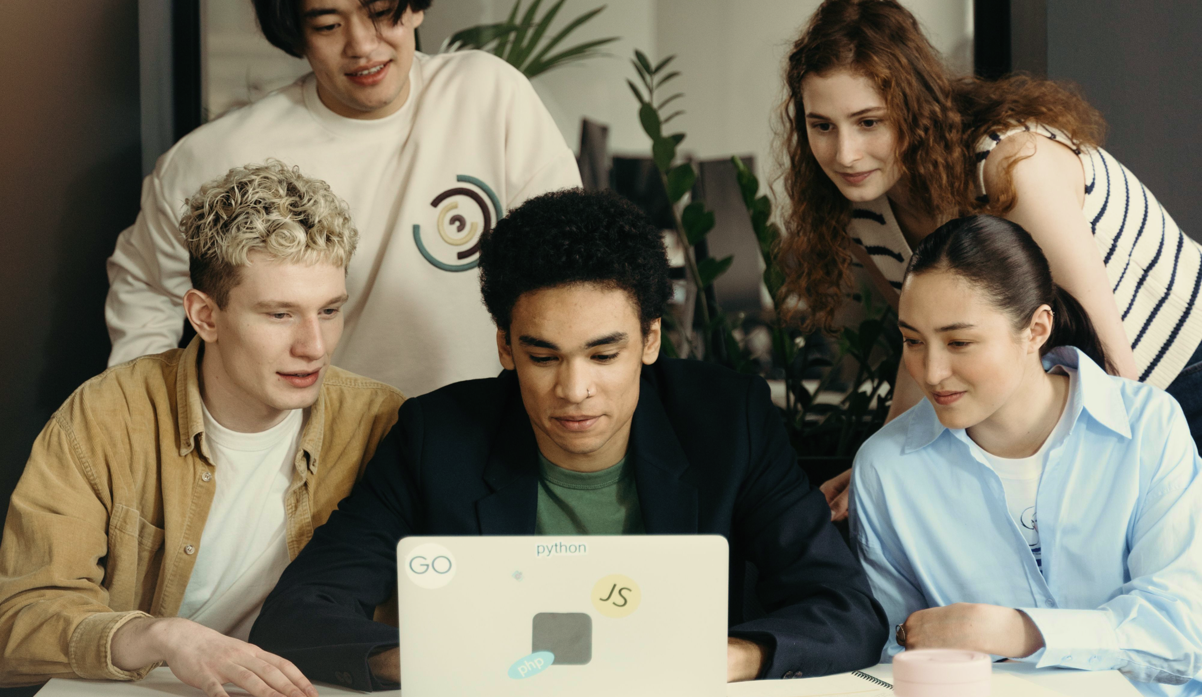Five young adults gather around a laptop covered with programming stickers, collaborating and smiling in a modern, plant-filled workspace.