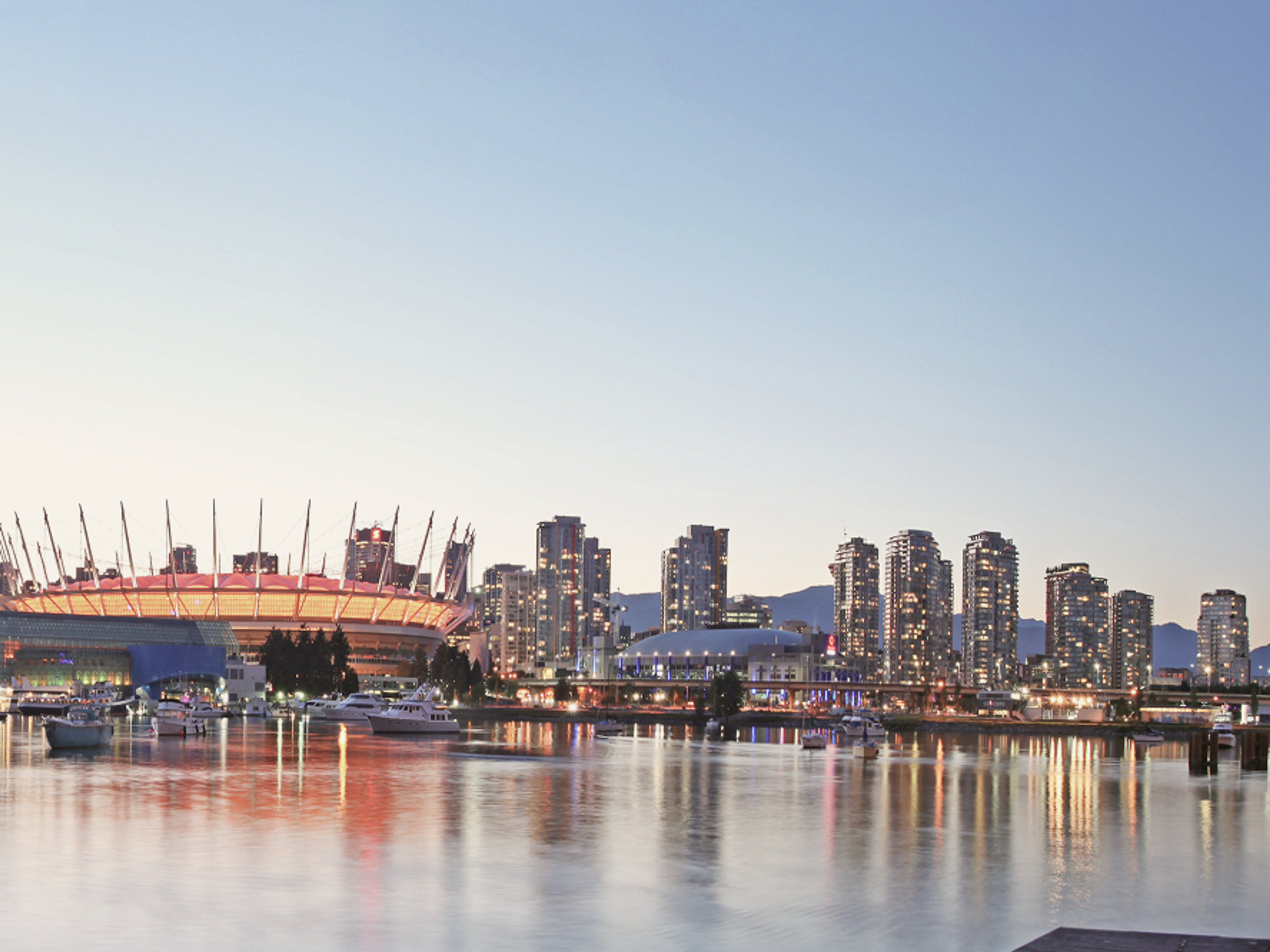 City skyline at dusk with a stadium lit in orange on the left, high-rise buildings reflected in calm water, boats docked along the waterfront, and mountains faintly visible in the background.