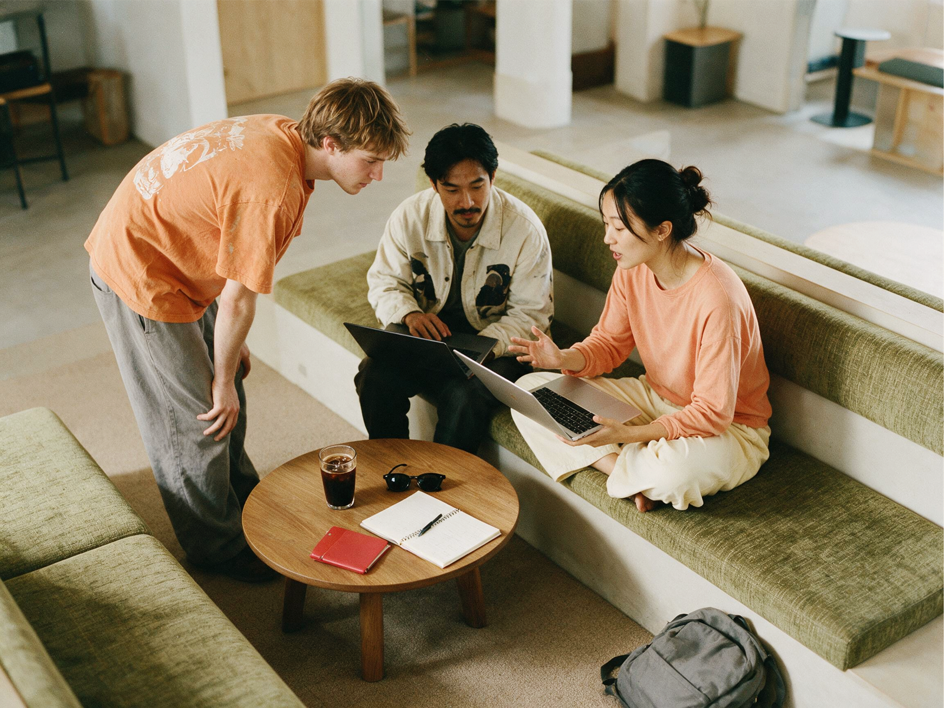 Three people are gathered around a coffee table with laptops, notebooks, and a drink, collaborating in a modern, casual workspace with green couches and soft lighting.