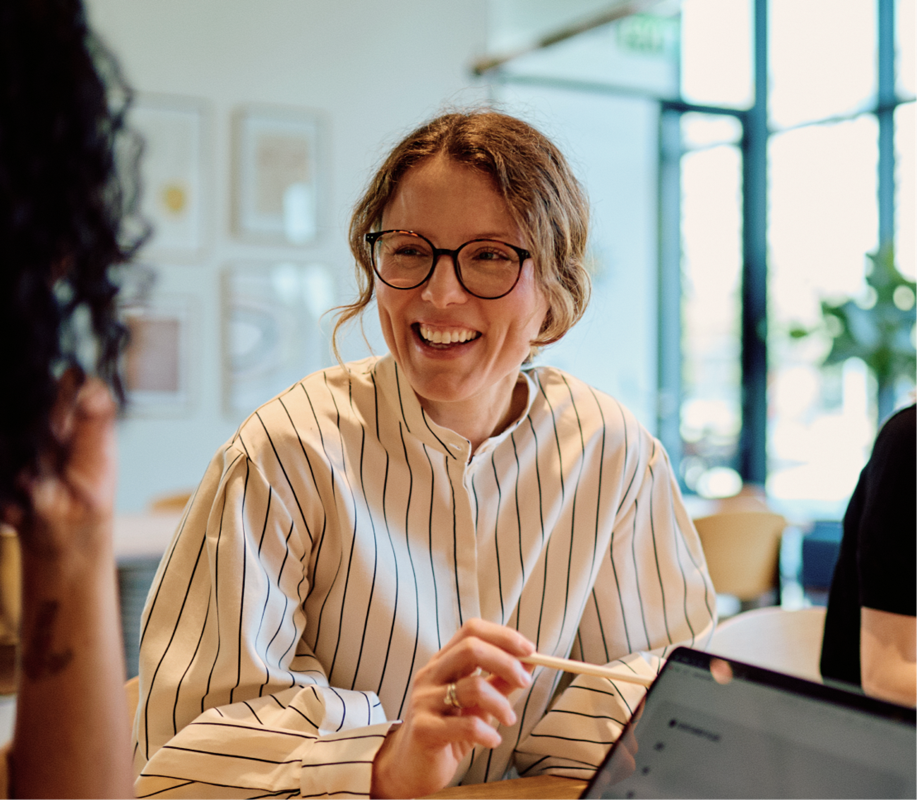 A woman wearing glasses and a striped shirt smiles while sitting at a table, holding a pen. She is engaged in conversation with another person in a brightly lit, modern office setting.
