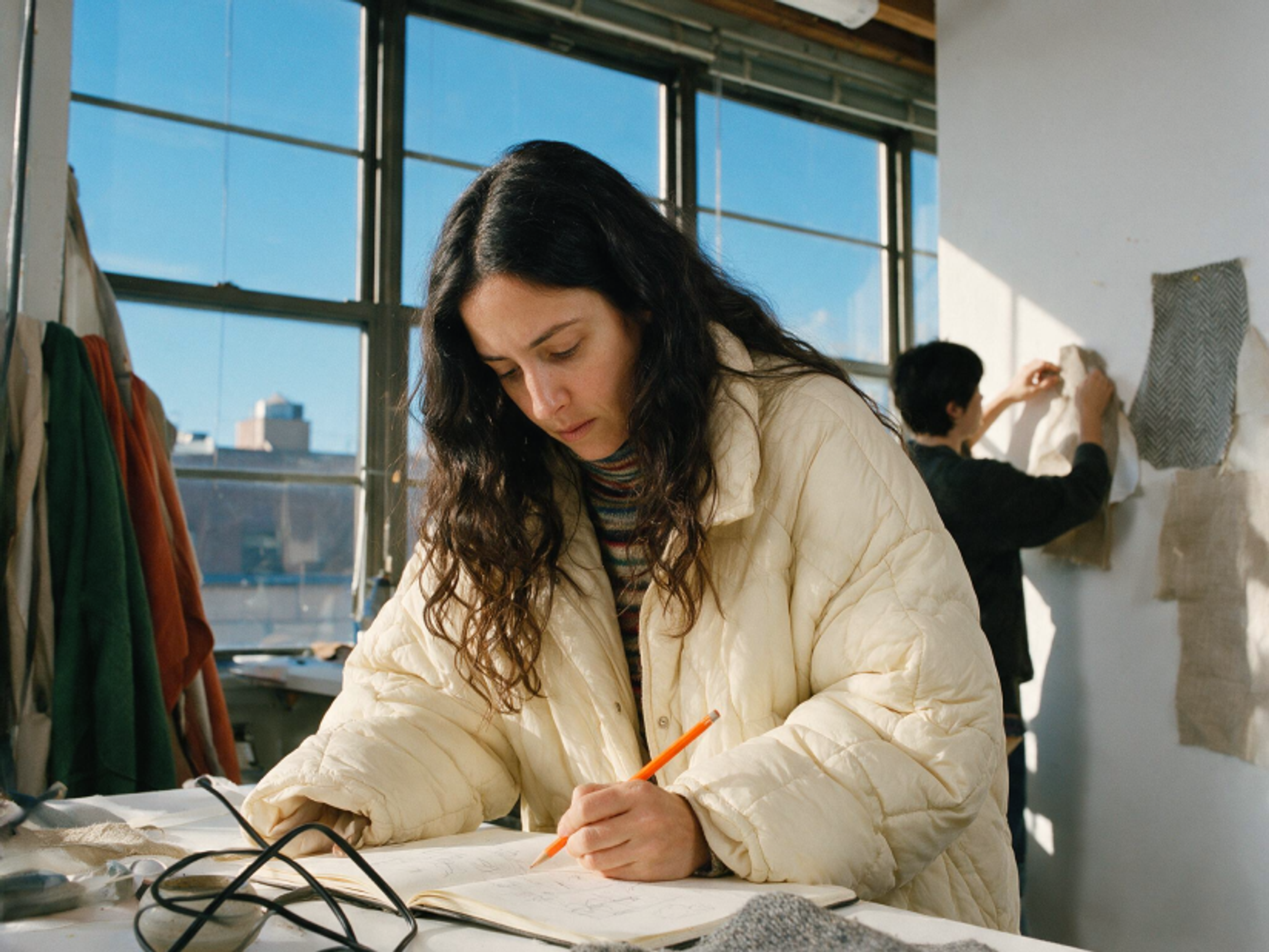 A person in a puffy cream jacket writes in a notebook at a sunlit table, while another person in the background hangs fabric samples on a white wall. Large windows show a cityscape outside.
