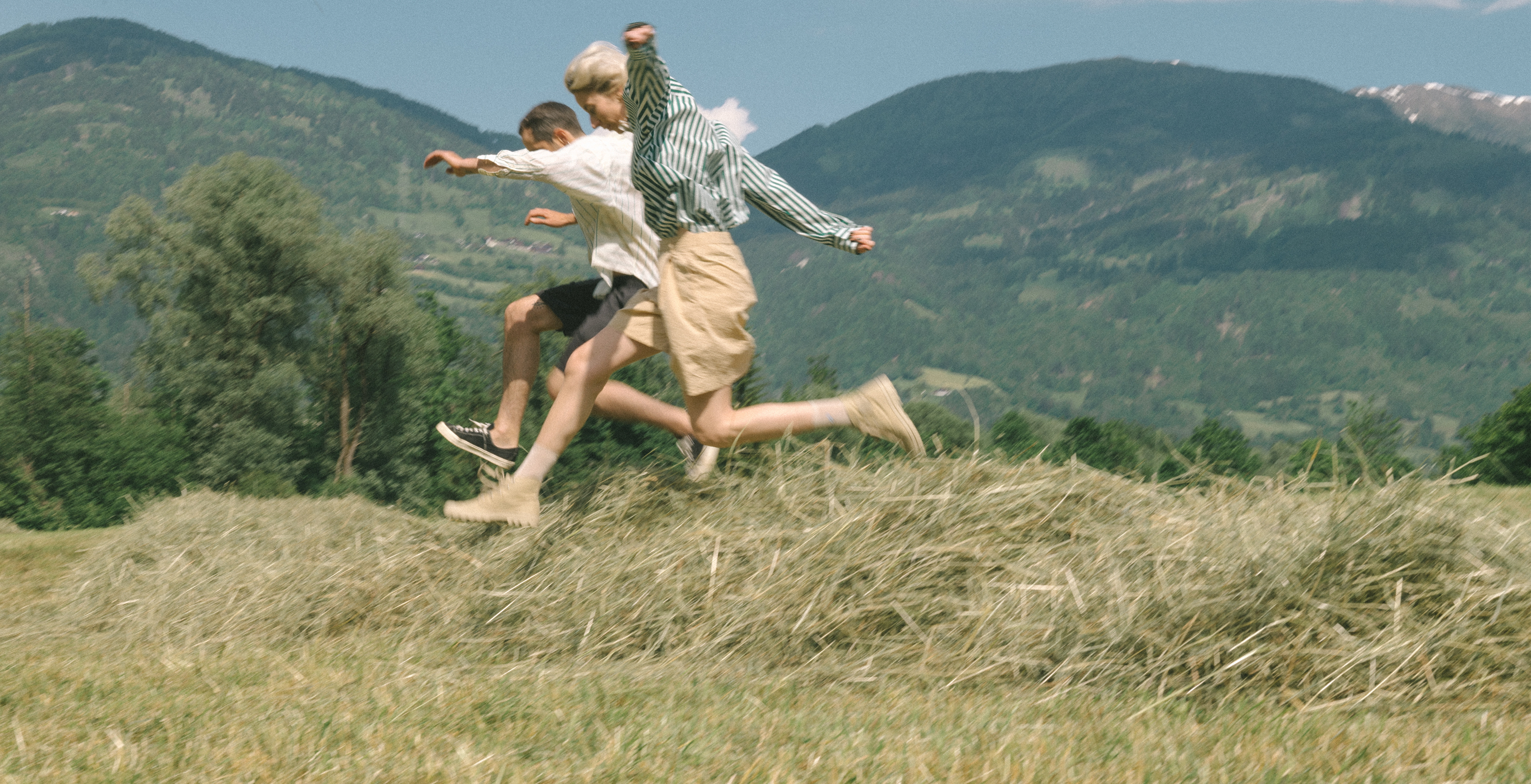 Two people are jumping over a pile of hay in a grassy field, with green trees and distant mountains in the background under a clear blue sky.