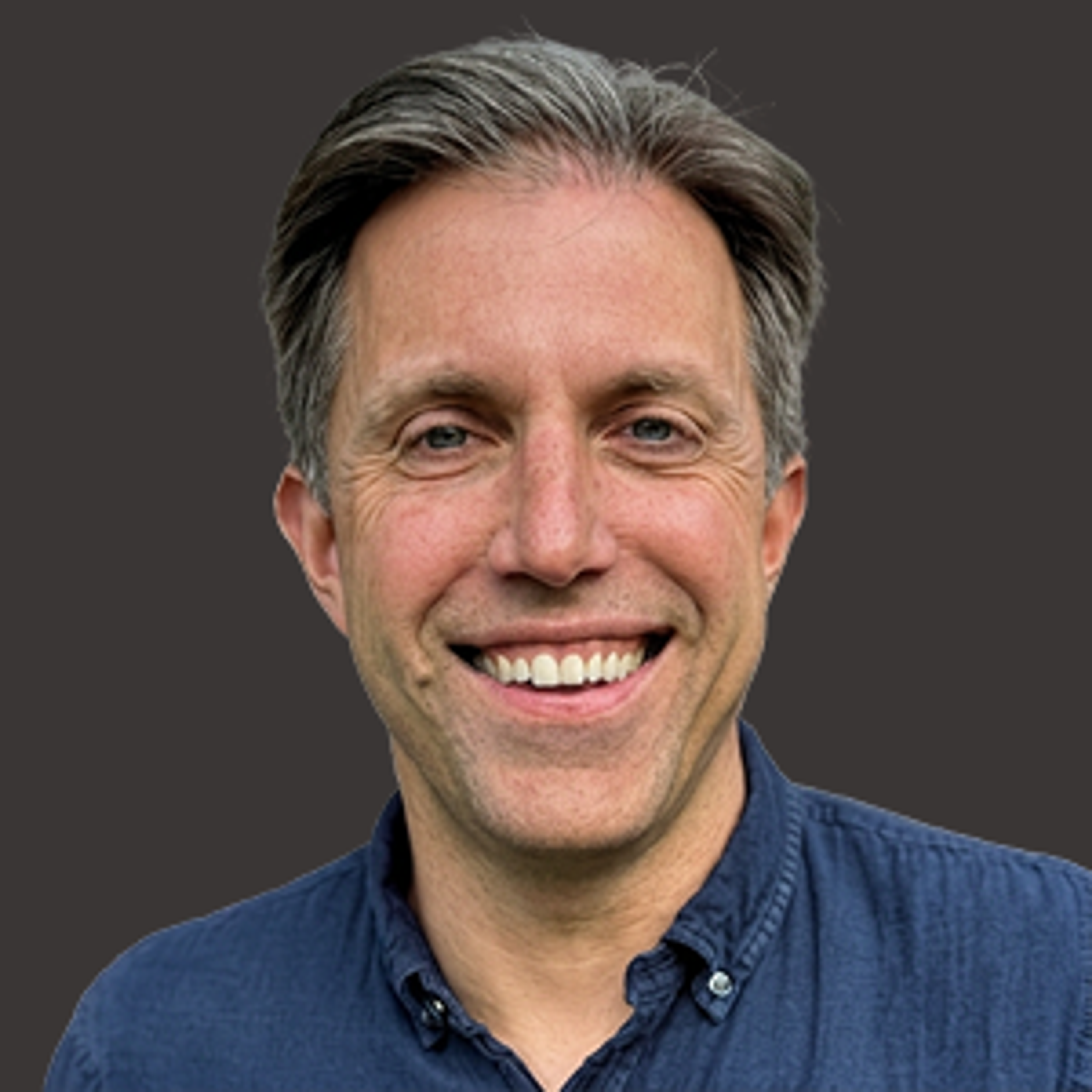 Smiling man with short grayish-brown hair wearing a dark blue button-up shirt, standing against a plain dark background.