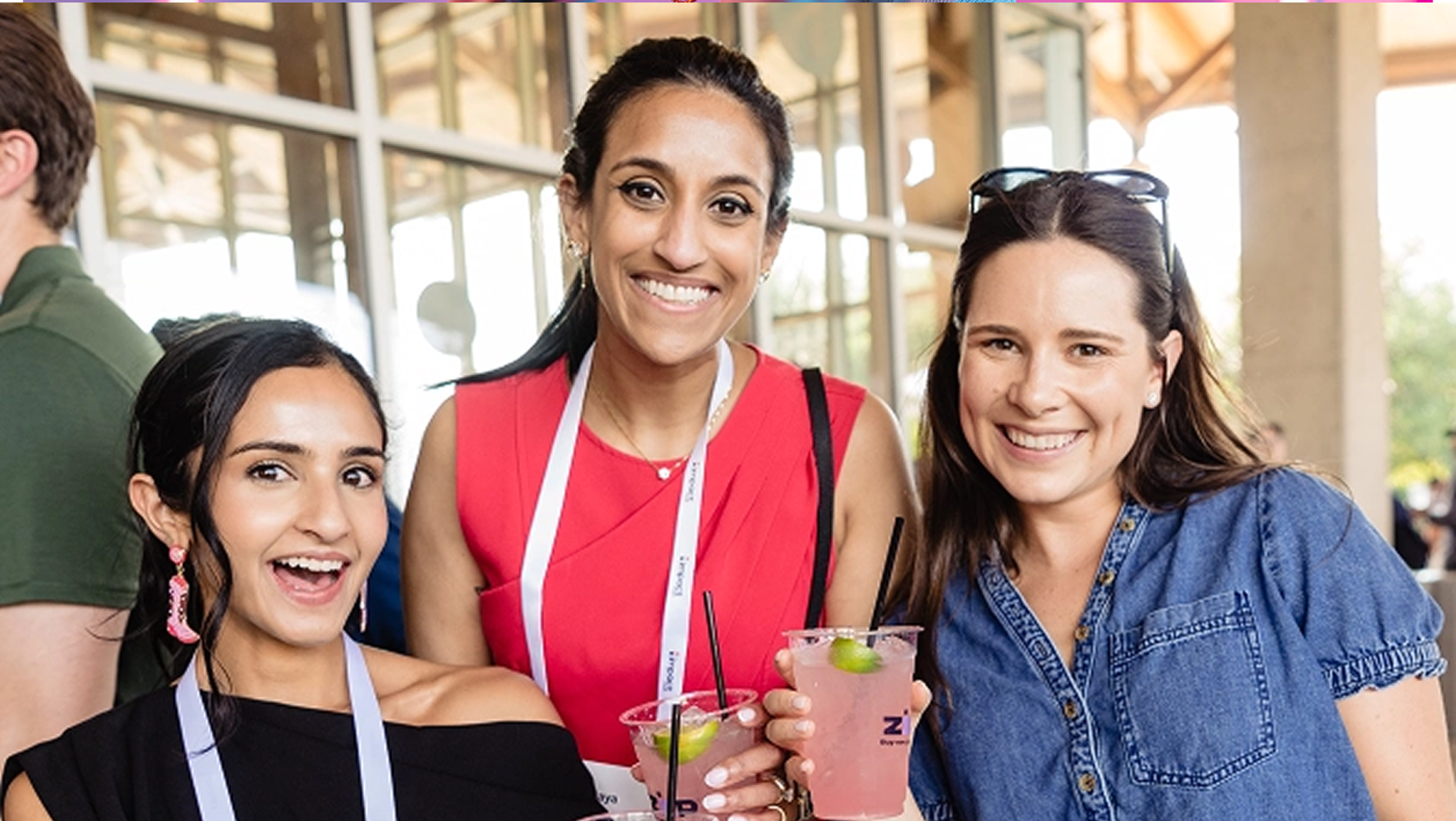 Three women smiling and holding pink drinks with lime slices at an outdoor event. They are wearing name lanyards and casual summer outfits, enjoying a cheerful moment together.