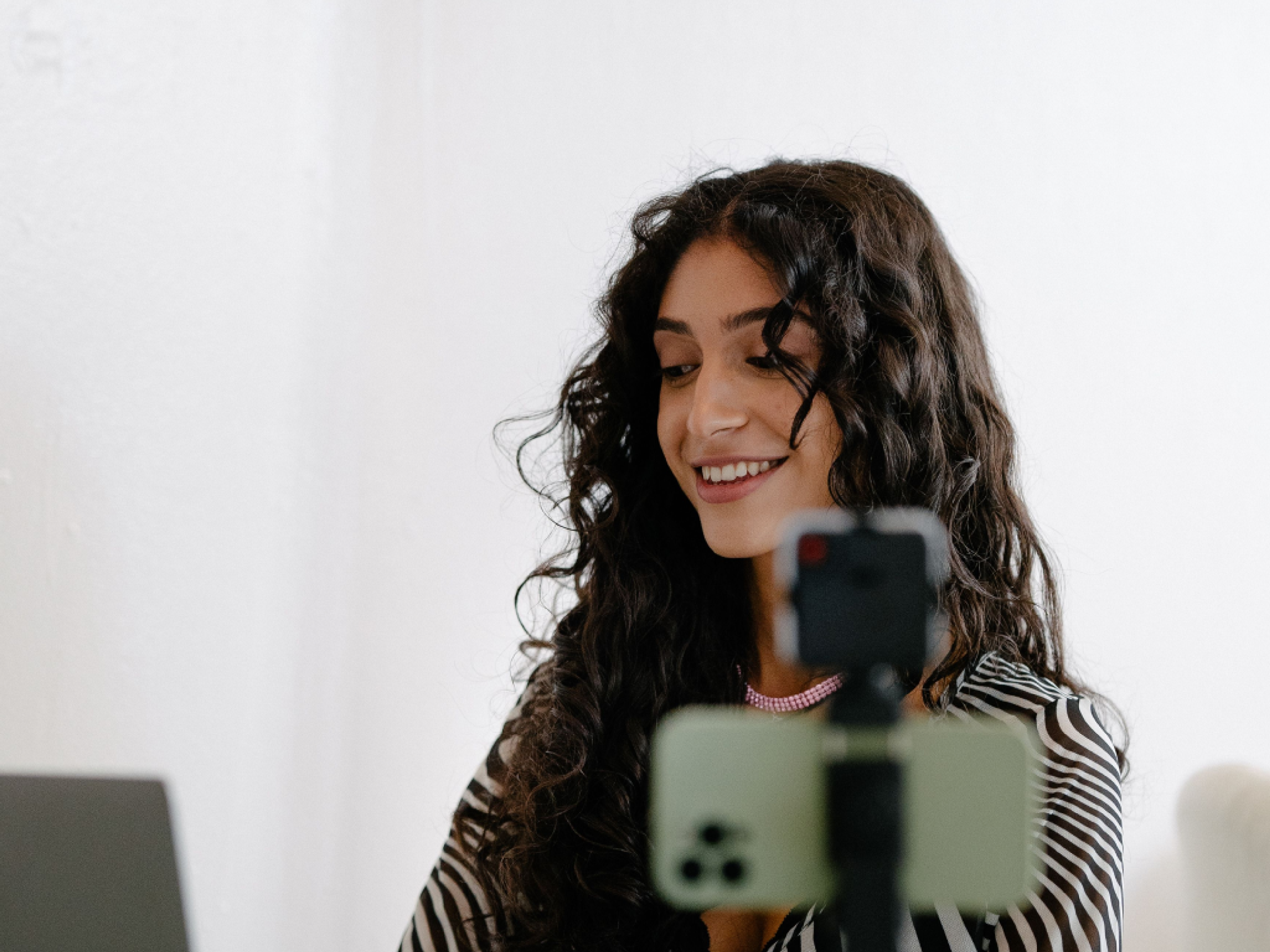 A woman with long curly hair smiles while looking at a laptop. A smartphone on a tripod is set up in front of her, suggesting she is recording or live streaming in a bright, indoor setting.