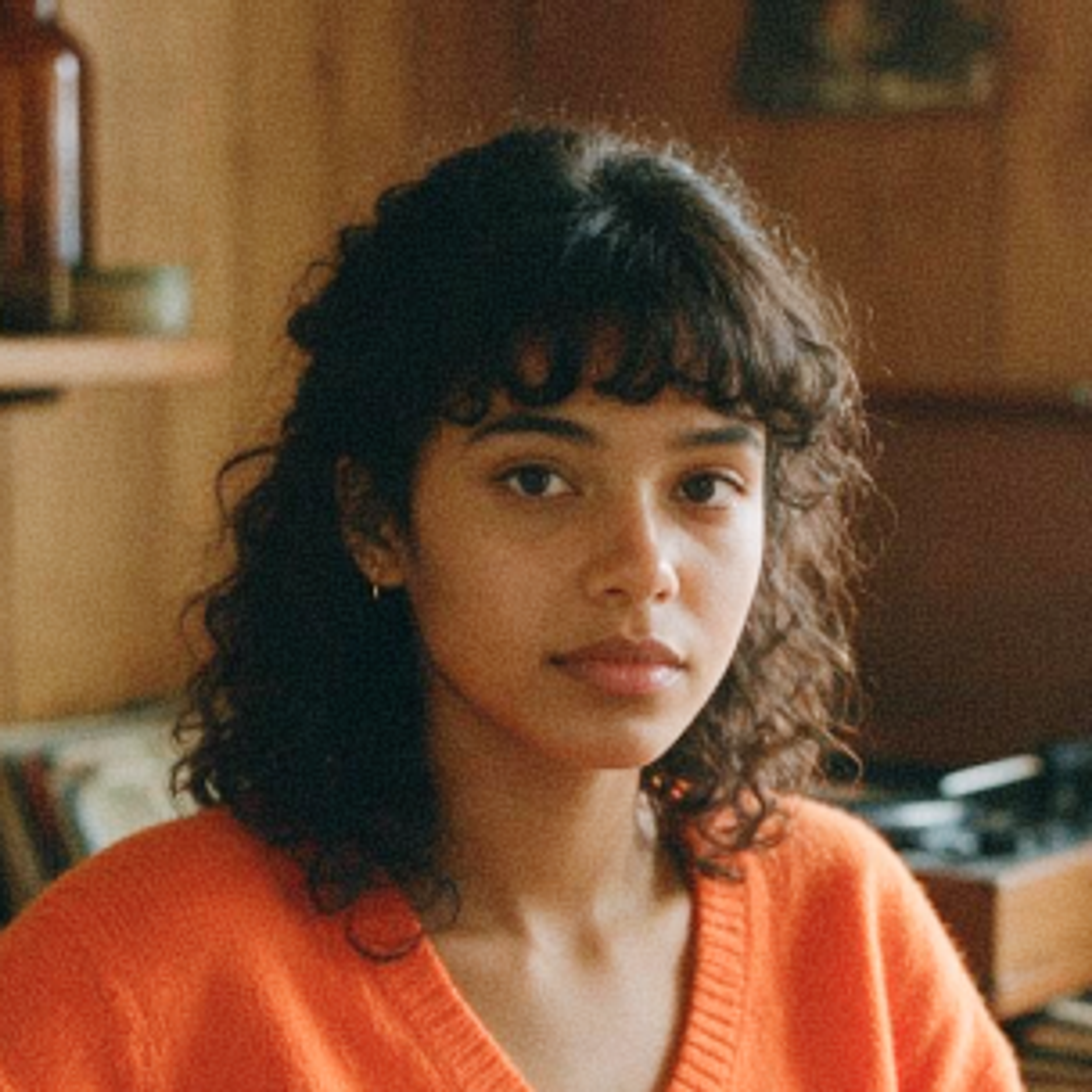 A young woman with curly dark hair and bangs, wearing a bright orange sweater, sits indoors with a neutral expression; wooden shelves and a record player are in the background.