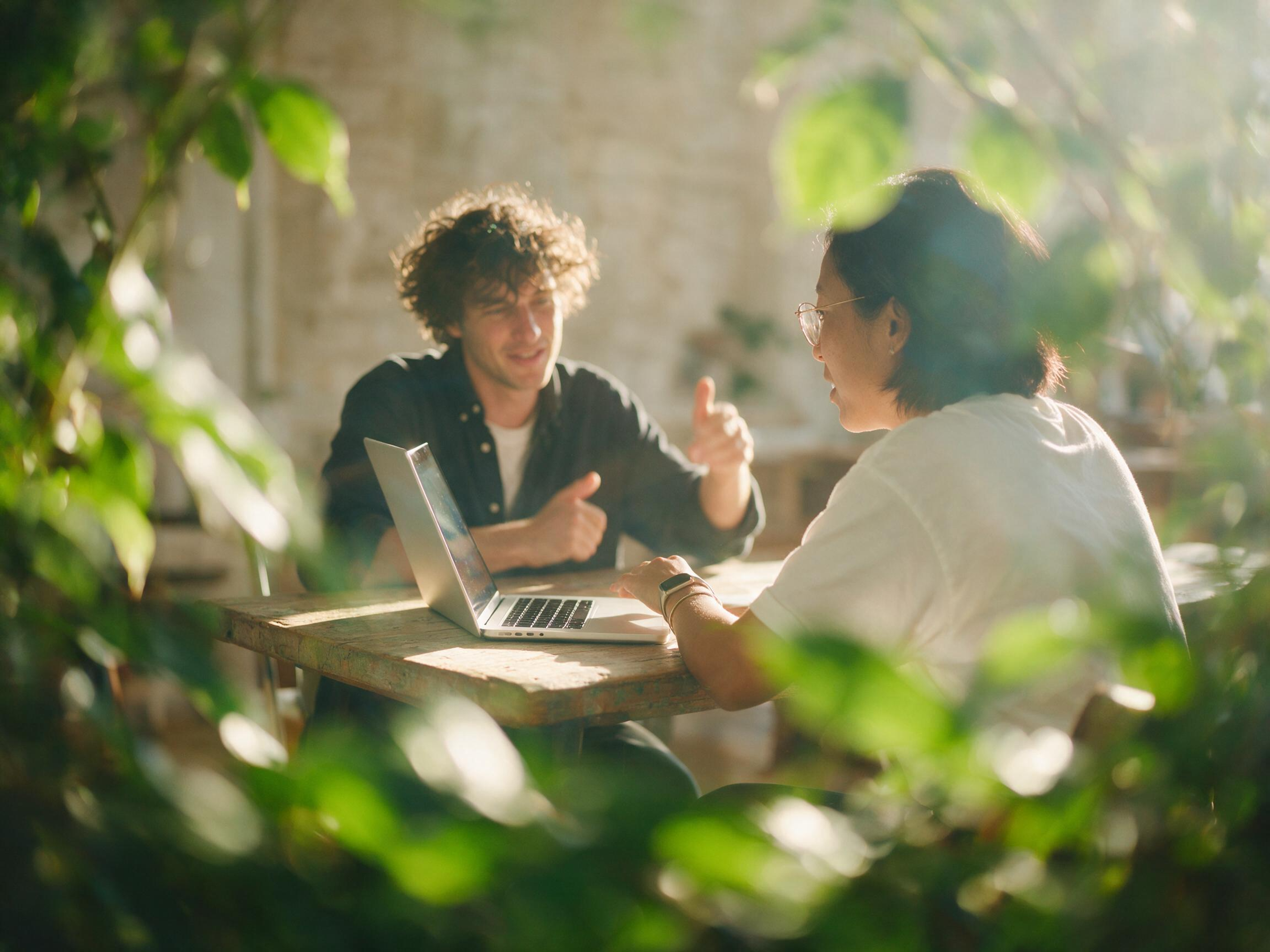 Two people sit at a wooden table with a laptop, having a conversation in a sunlit, plant-filled room. Sunlight filters through green leaves, creating a warm and relaxed atmosphere.