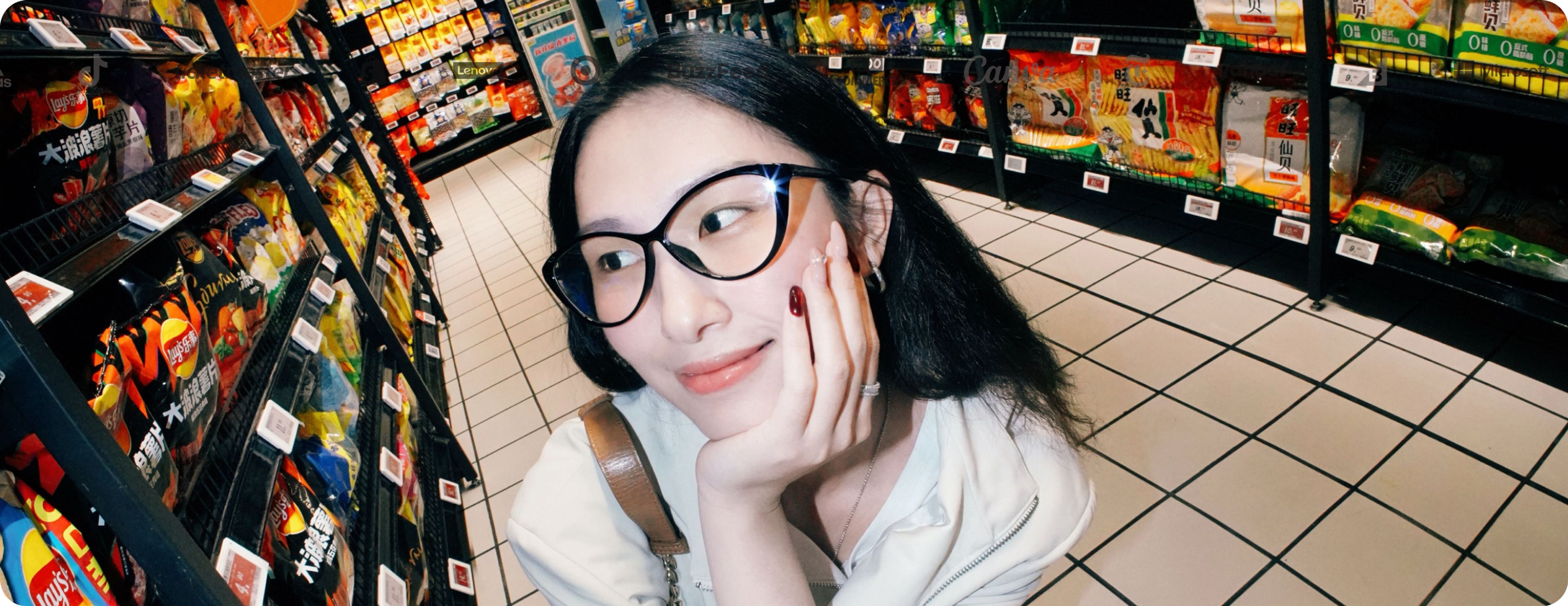A person with glasses smiles while standing in an aisle of a grocery store, surrounded by colorful snacks on the shelves.
