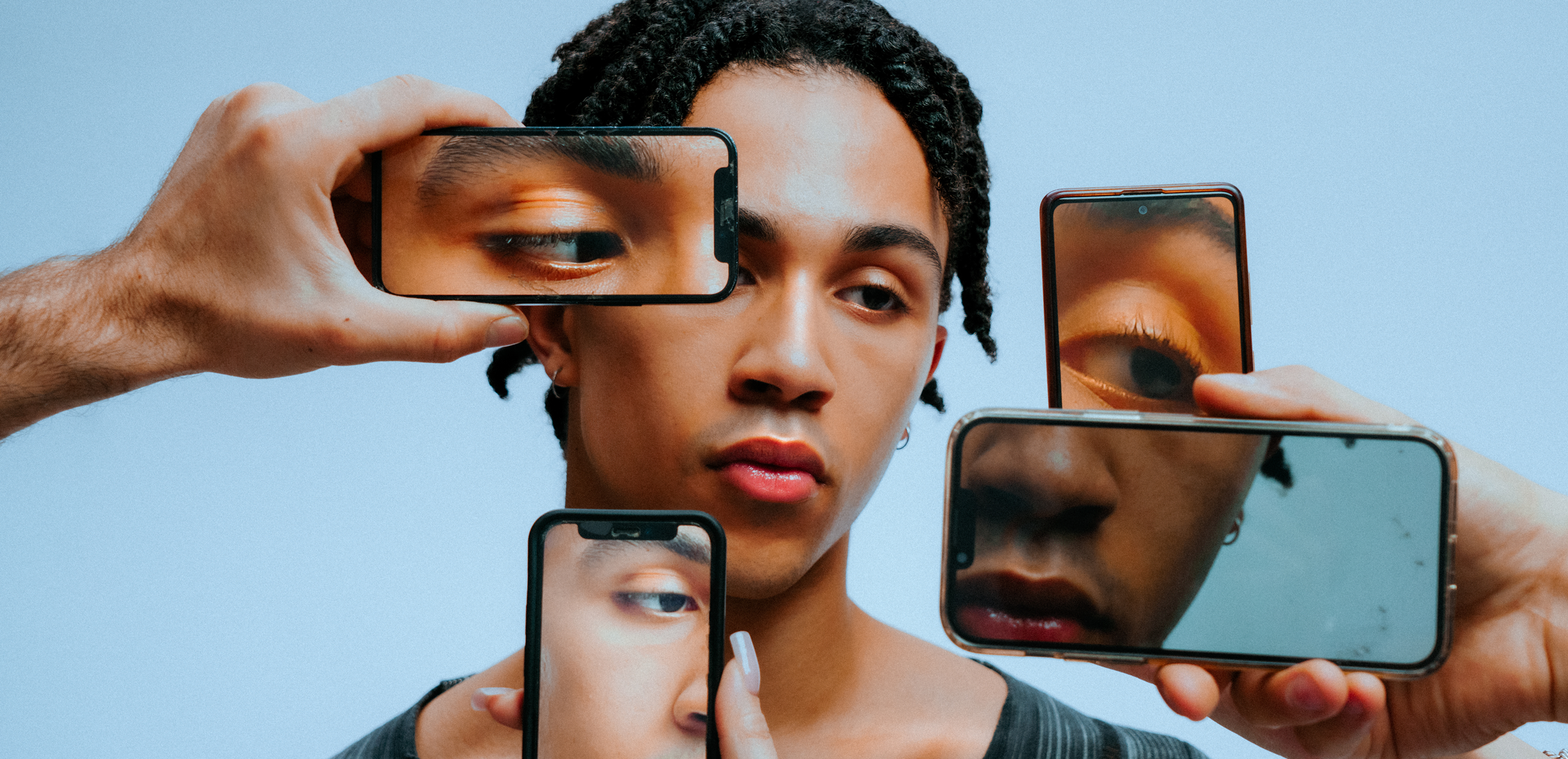 A person with braided hair looks forward as hands hold phones showing close-ups of their eyes, lips, and face around them, set against a plain background.