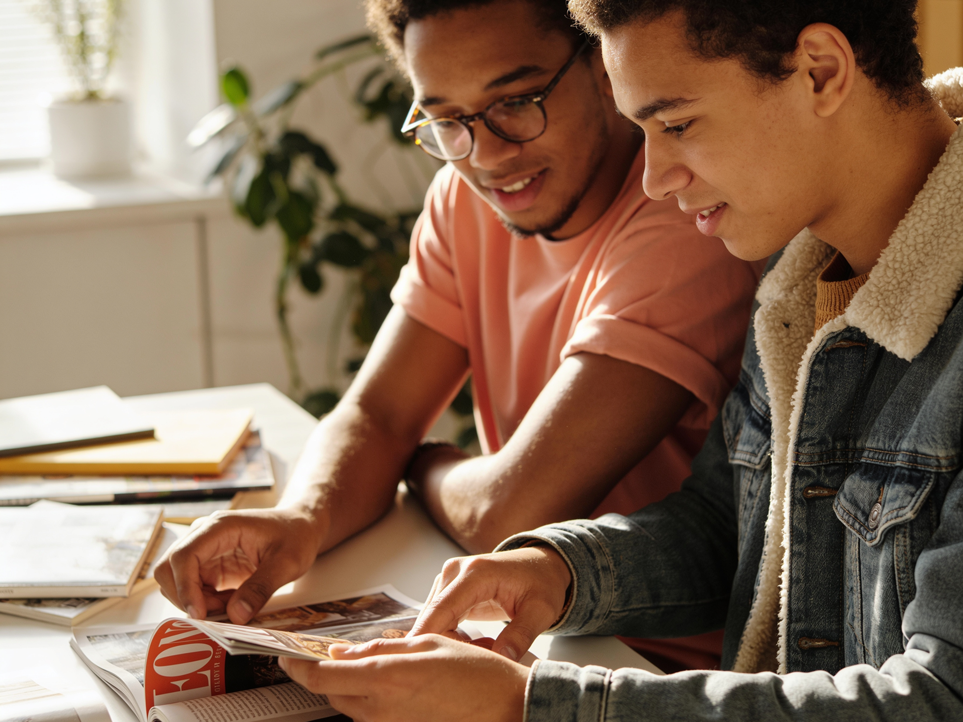 Two young men sit at a table, smiling and looking at a magazine together. Sunlight streams through a window, and books and papers are spread around them. A green potted plant is visible in the background.