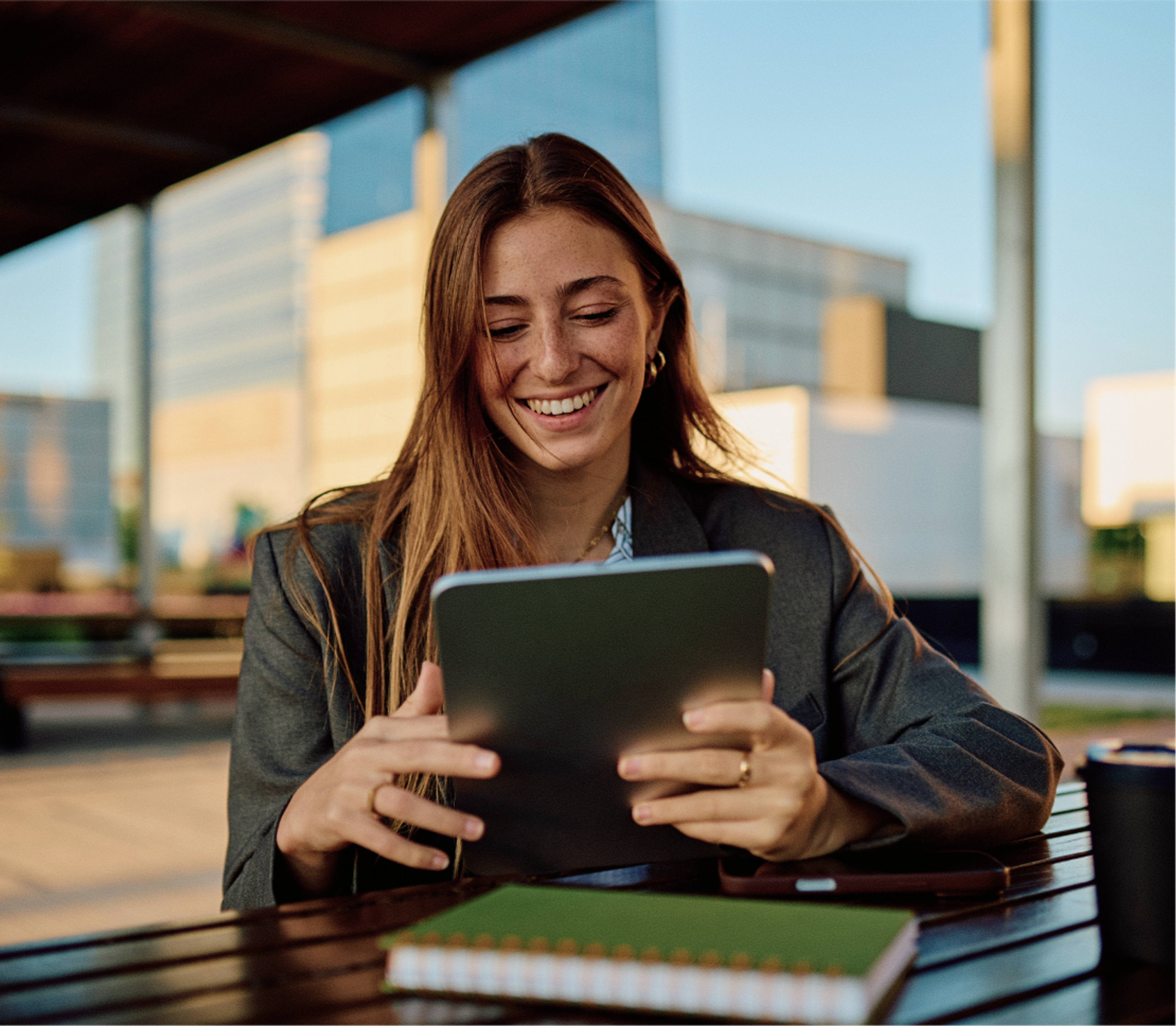 A woman with long brown hair smiles while using a tablet outdoors at a table. She is wearing a blazer, and a closed green notebook lies in front of her. Modern buildings are visible in the background.