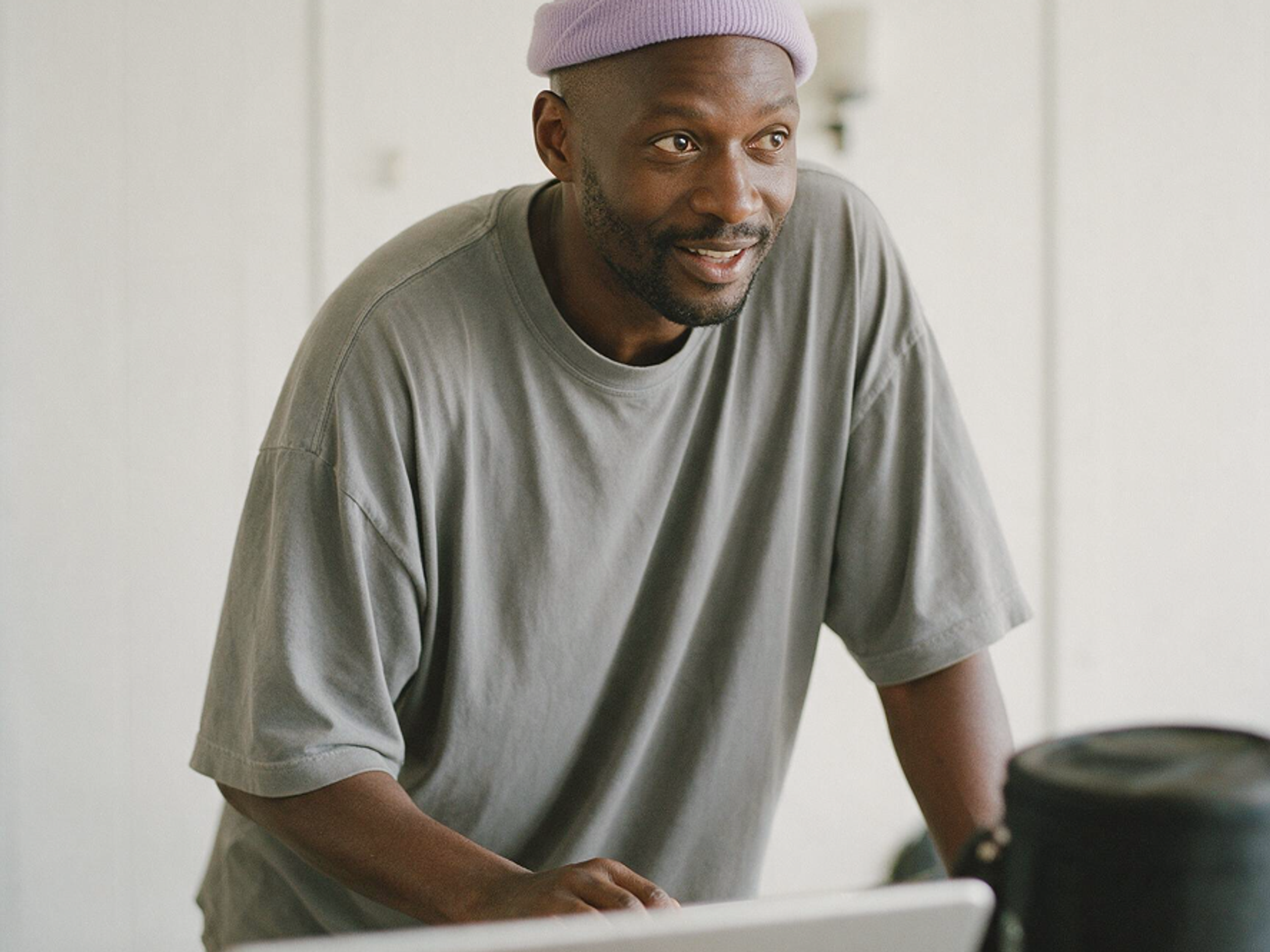 A man wearing a purple beanie and gray t-shirt leans forward and smiles, looking toward a laptop in a bright room.