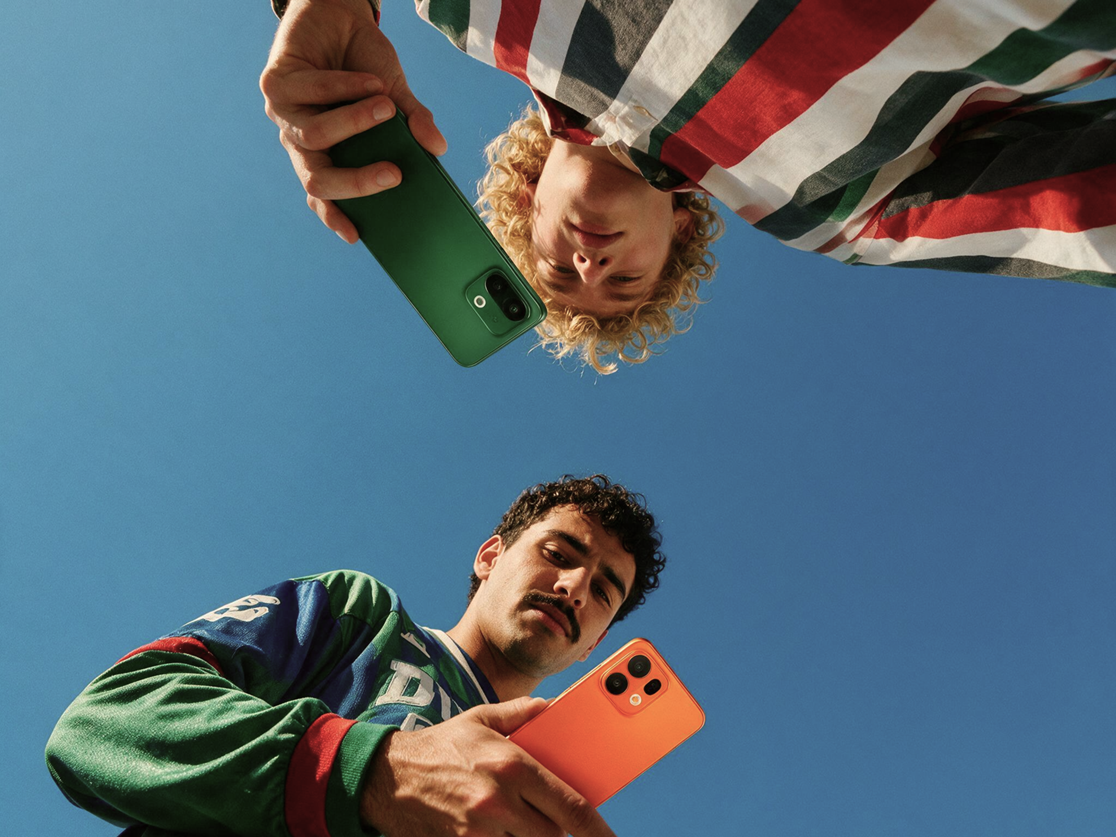 Two people are looking down at the camera, each holding a bright-colored smartphone, with a clear blue sky in the background.